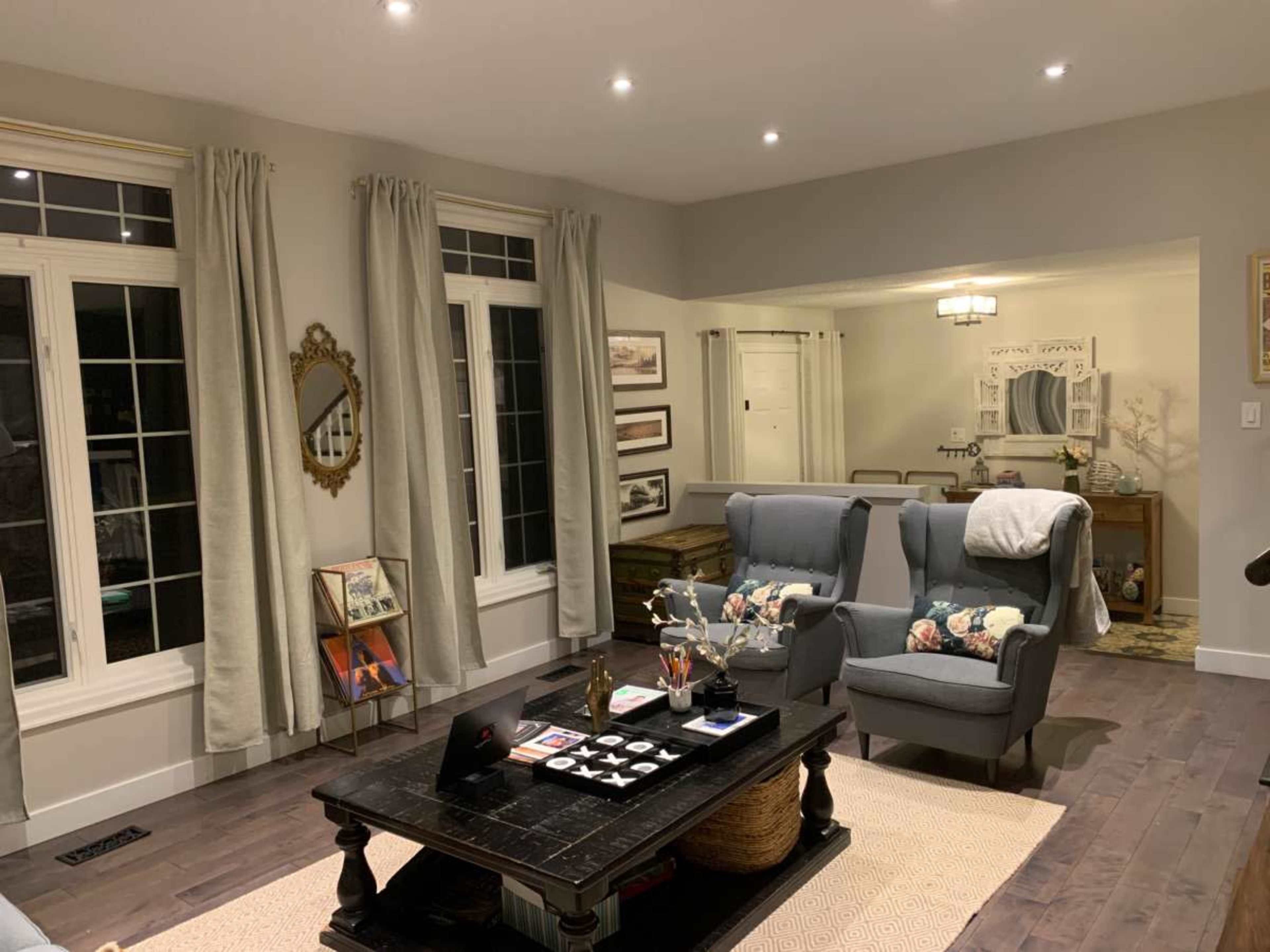The image shows a cozy living room with a dark wood coffee table, two light blue armchairs, large windows dressed with curtains, and framed artwork on the walls.