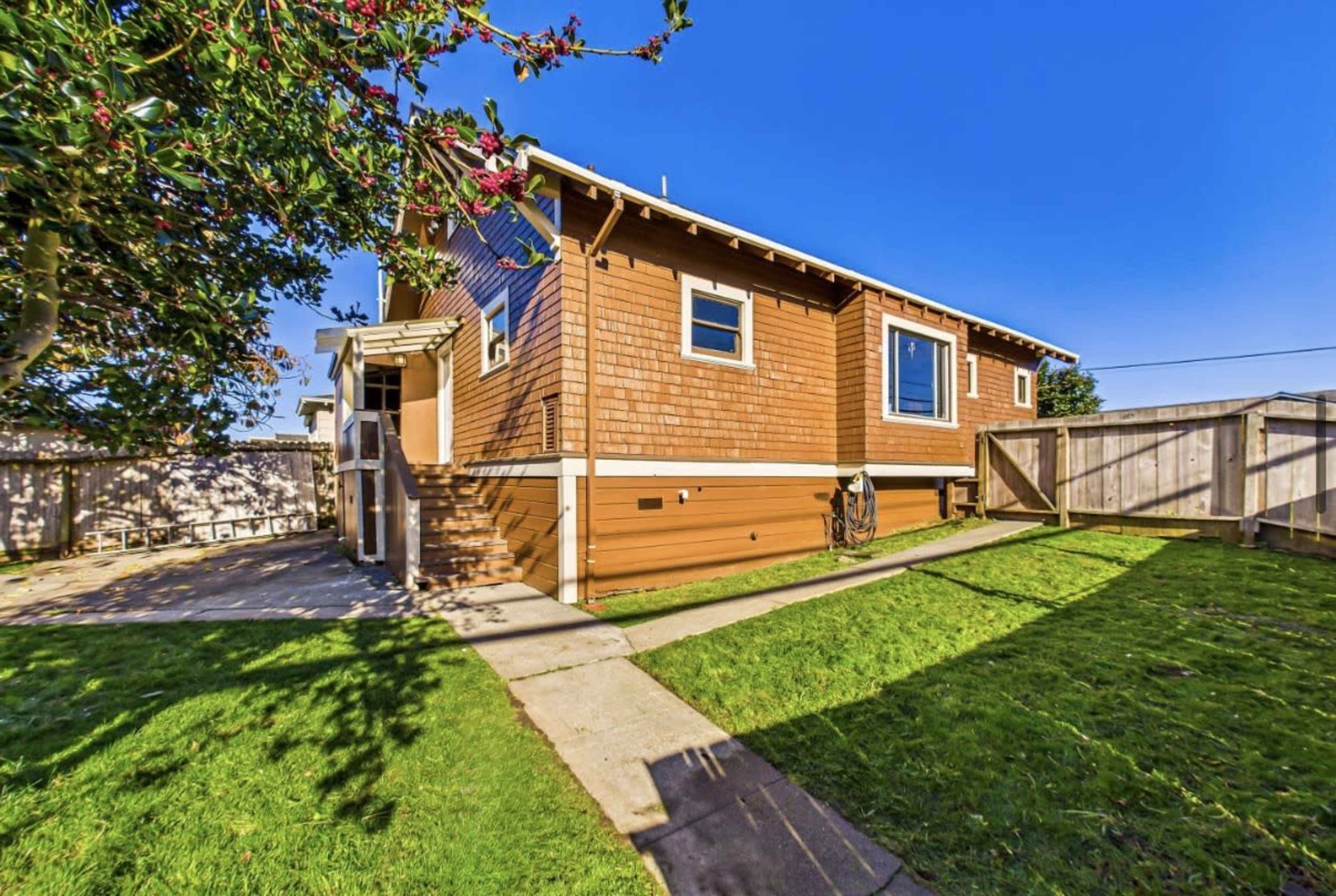 The image shows a side view of a single-story house with a grassy yard and paved pathway leading to the entrance.