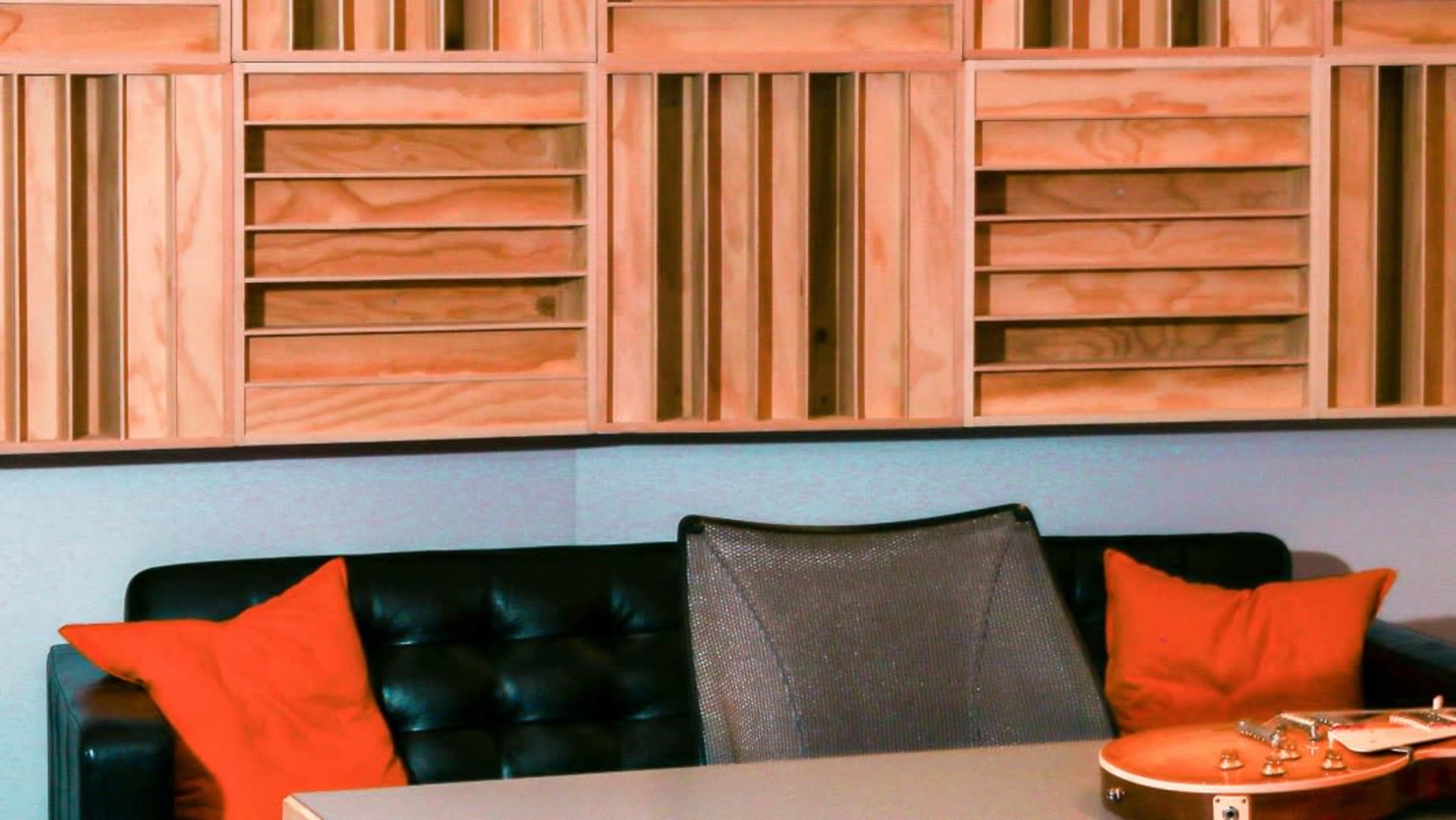 The image shows a modern workspace with a black couch adorned with orange cushions, a simple table, and a wooden partition featuring slatted design behind it.