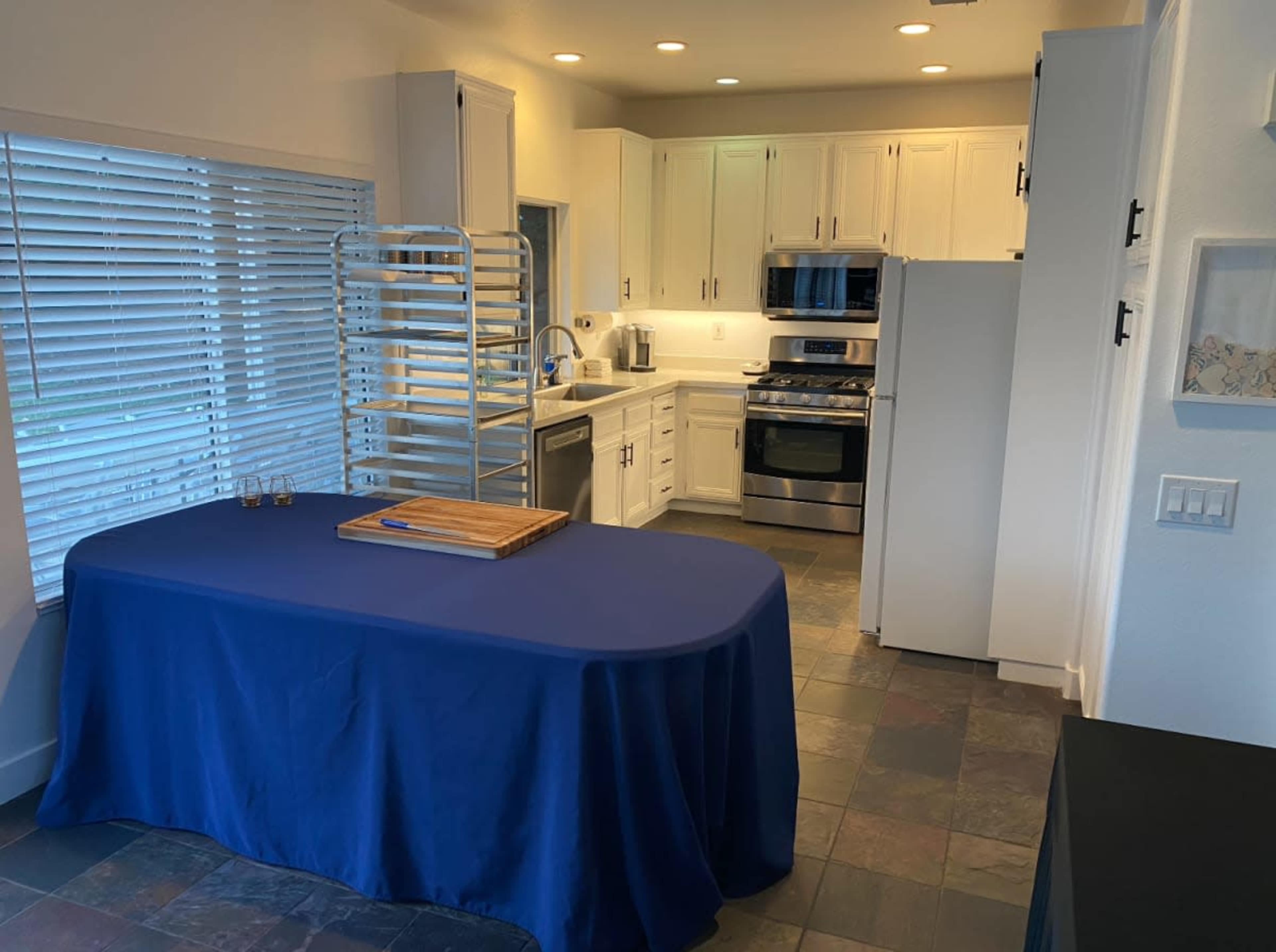 A clean kitchen features a blue tablecloth-covered table in the foreground, with stainless steel appliances and white cabinetry in the background.