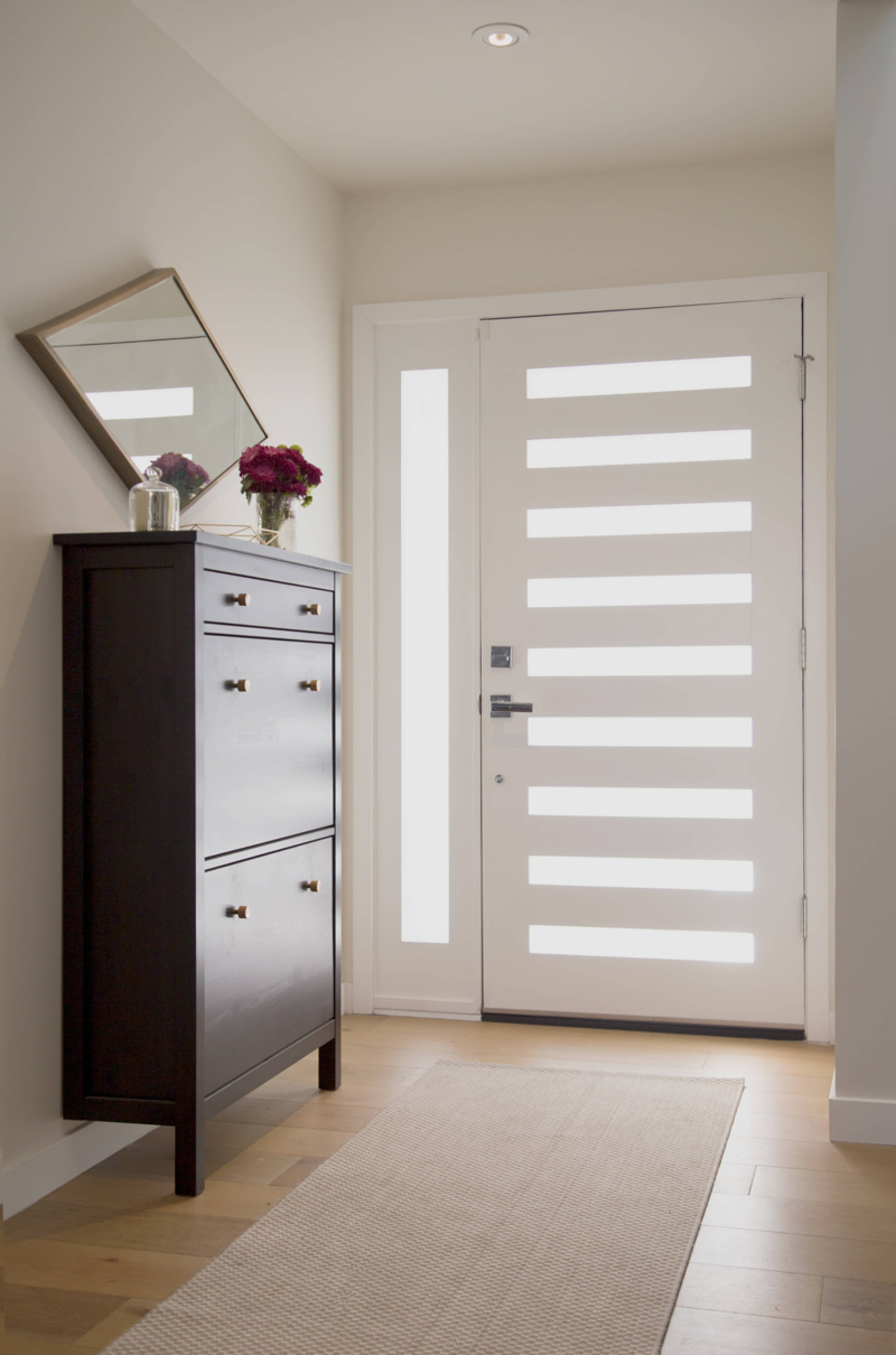 The image shows a modern entryway featuring a white striped door, a tall dark cabinet, a small mirror on the wall, and a flower vase on top of the cabinet.