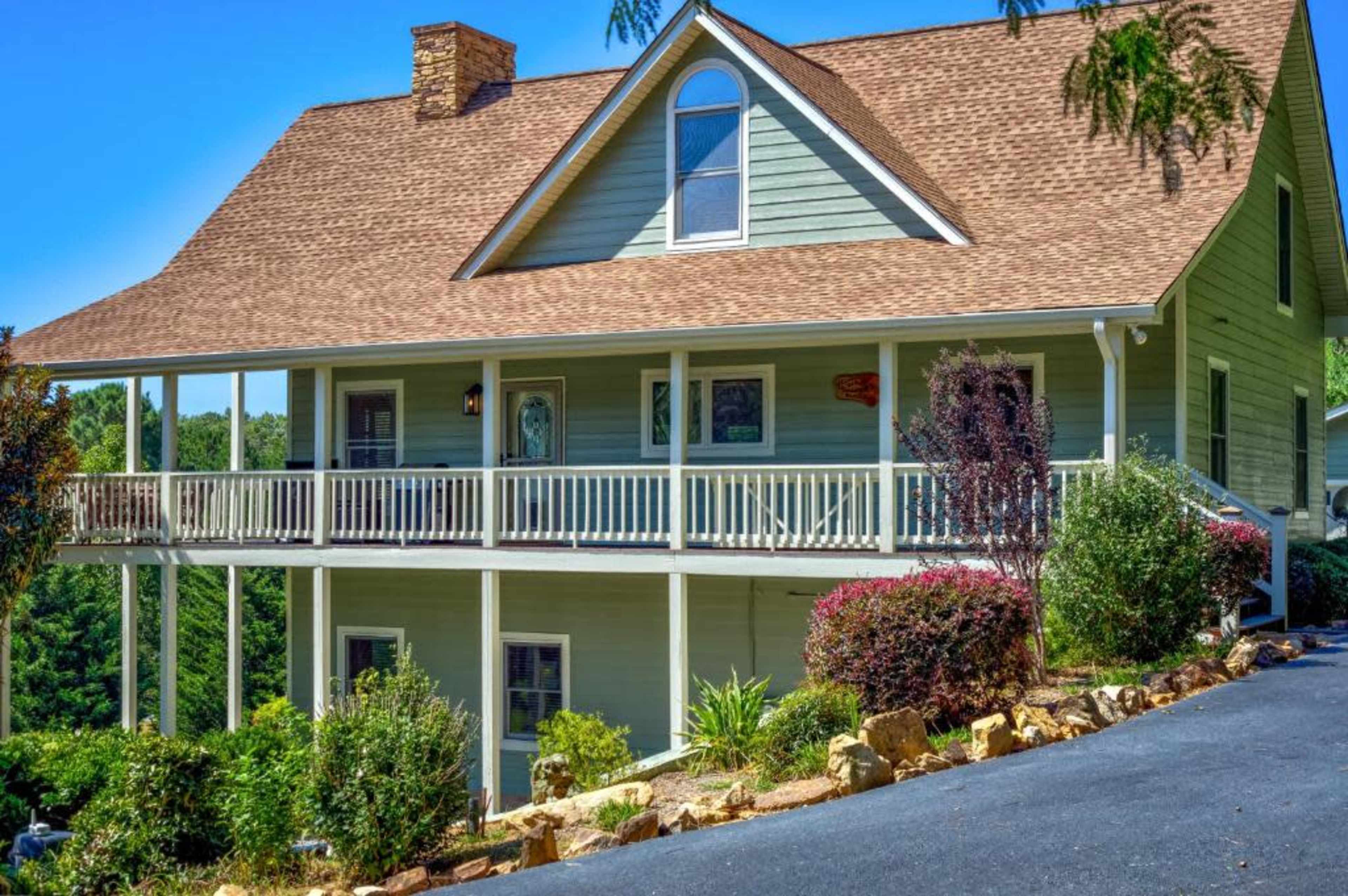 A two-story house with a sloped roof and a wraparound porch stands on a landscaped lot, surrounded by greenery and flowering shrubs.