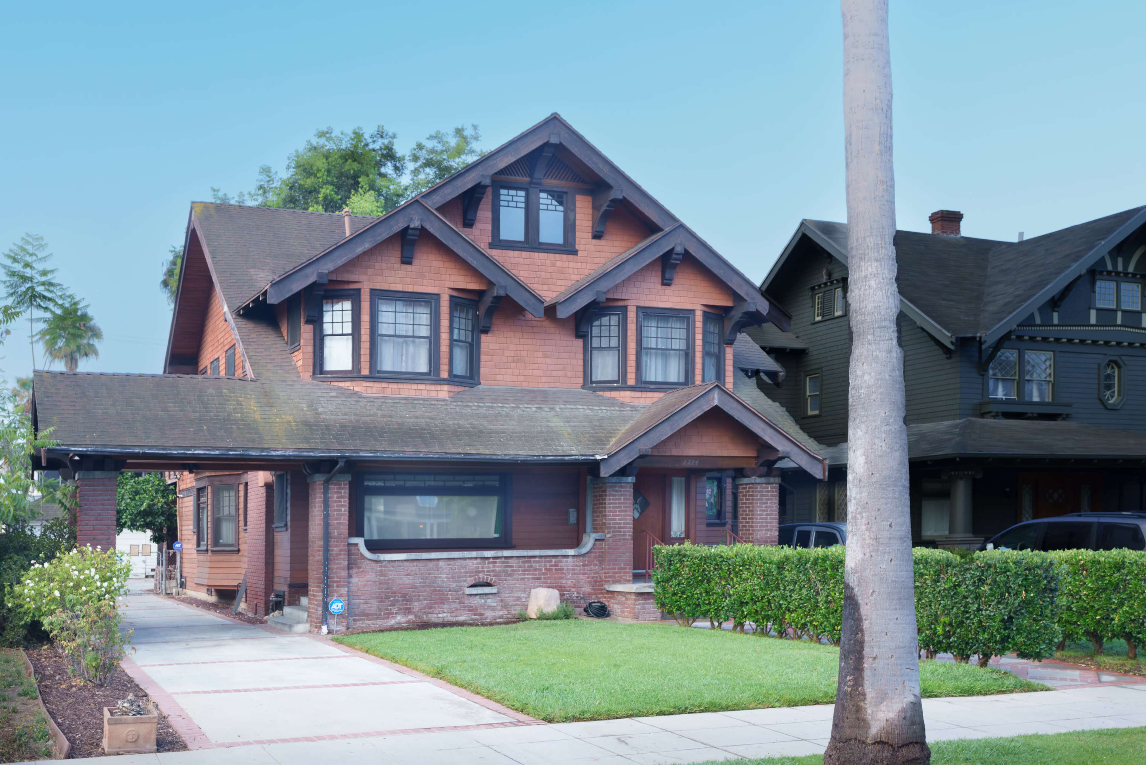 The image shows a two-story, wooden house with a steeply pitched roof and decorative gables, situated beside another similar-style house in a residential area.