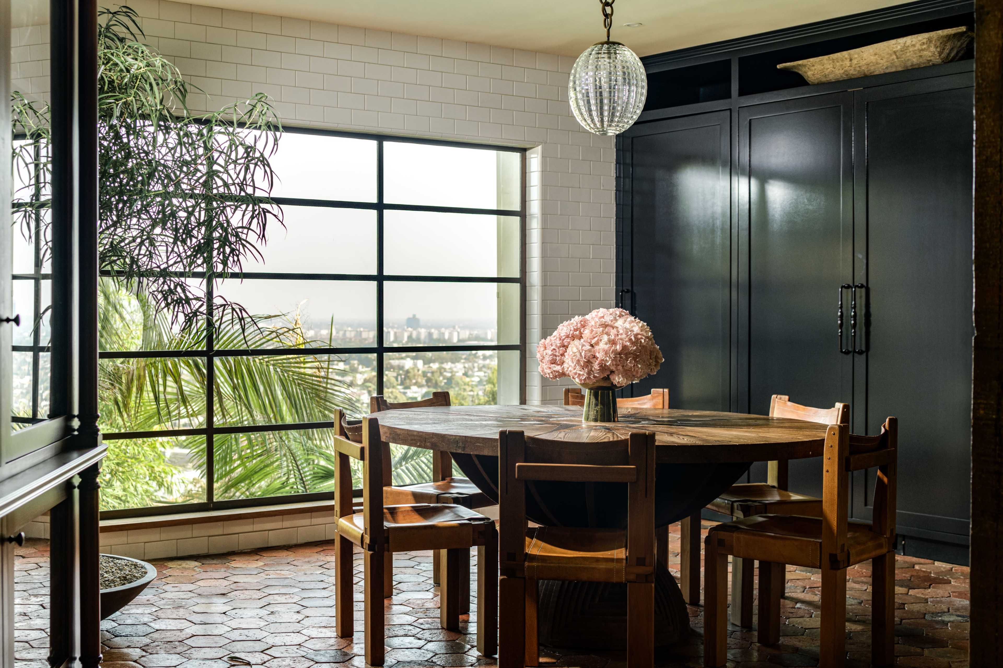 A round wooden dining table surrounded by wooden chairs is set near large windows displaying a view of greenery and a cityscape.