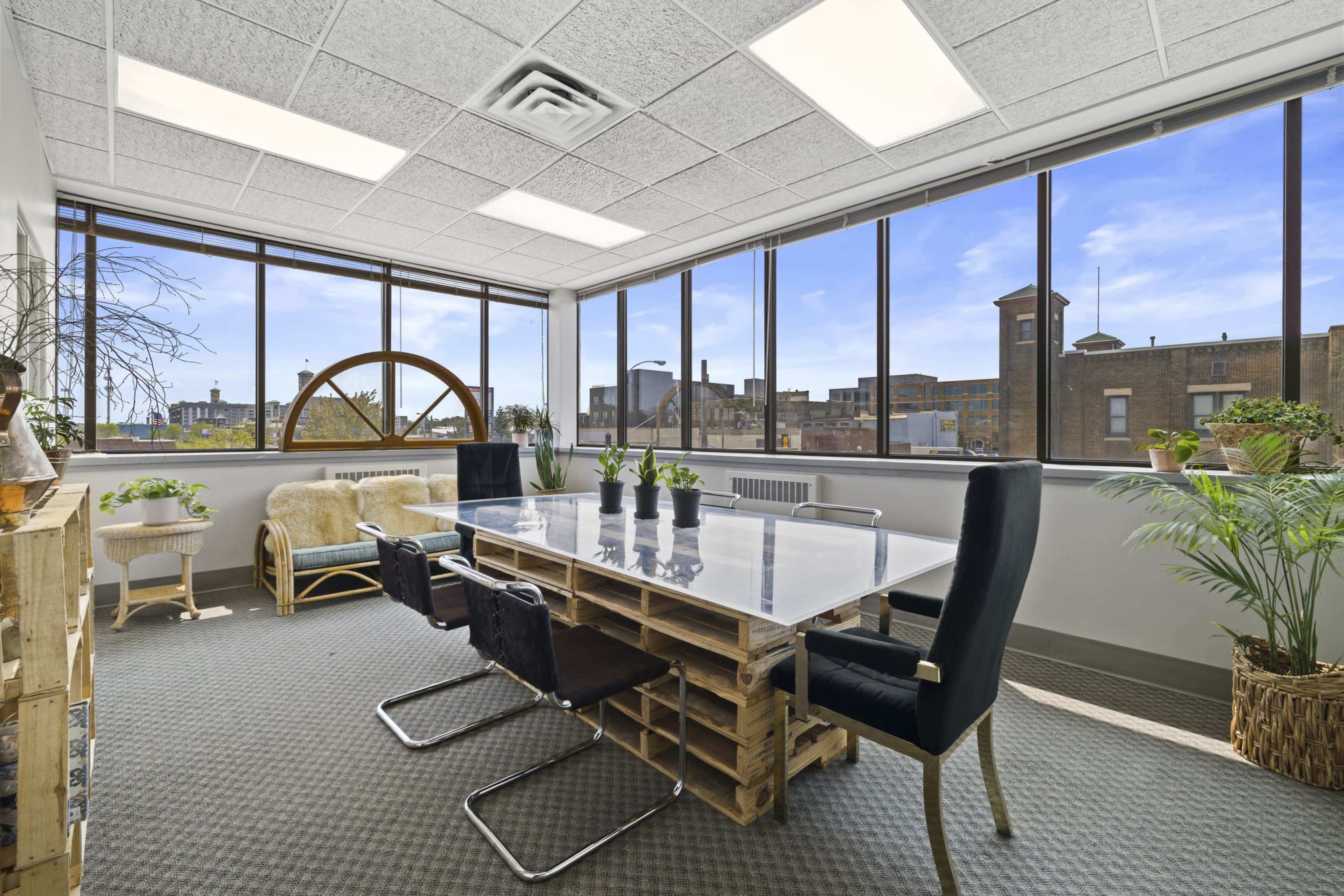 A bright office meeting room with large windows, featuring a glass table surrounded by black chairs and various plants.