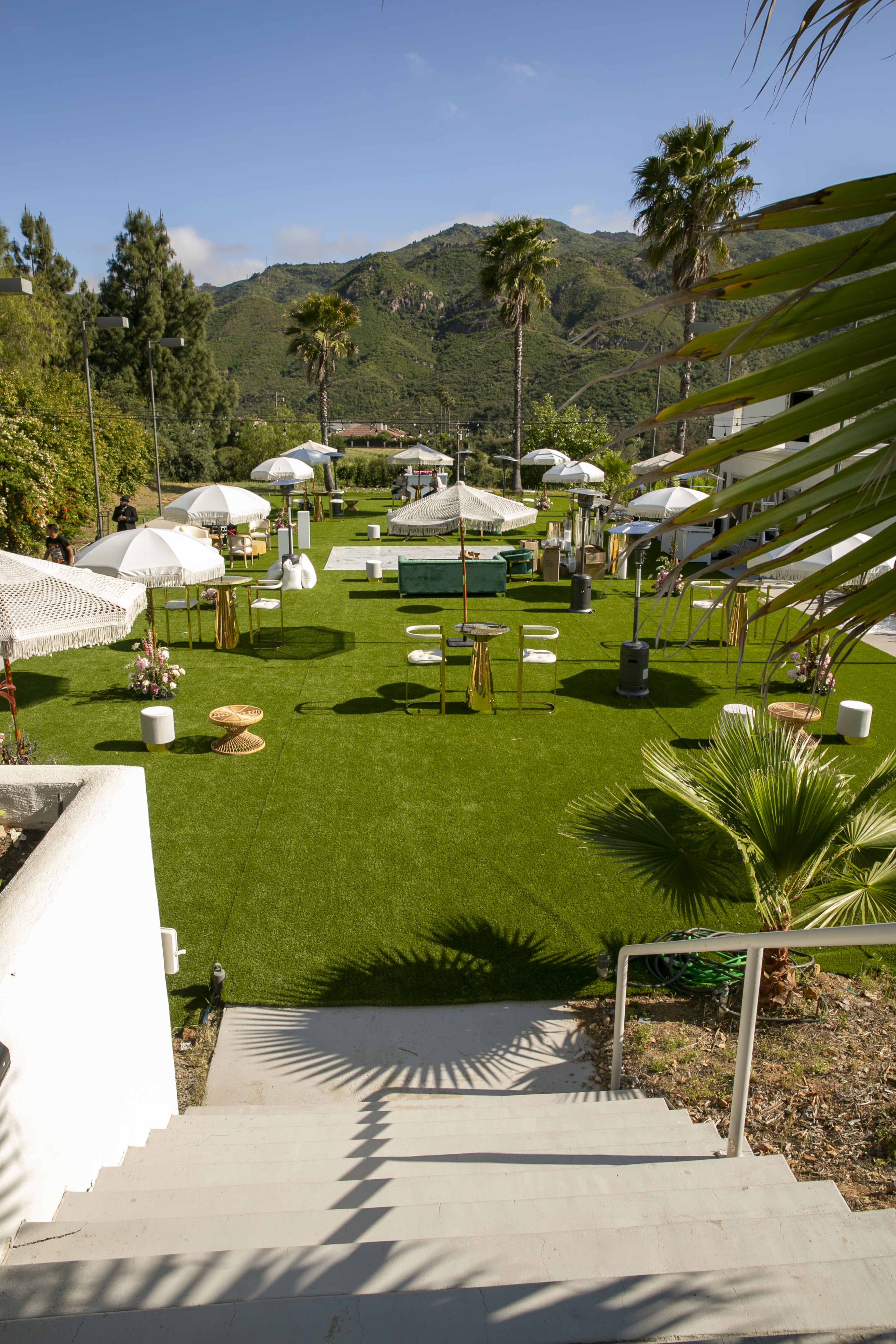 A landscaped outdoor area features green artificial grass, seating arrangements, and several large umbrellas, set against a backdrop of mountains.