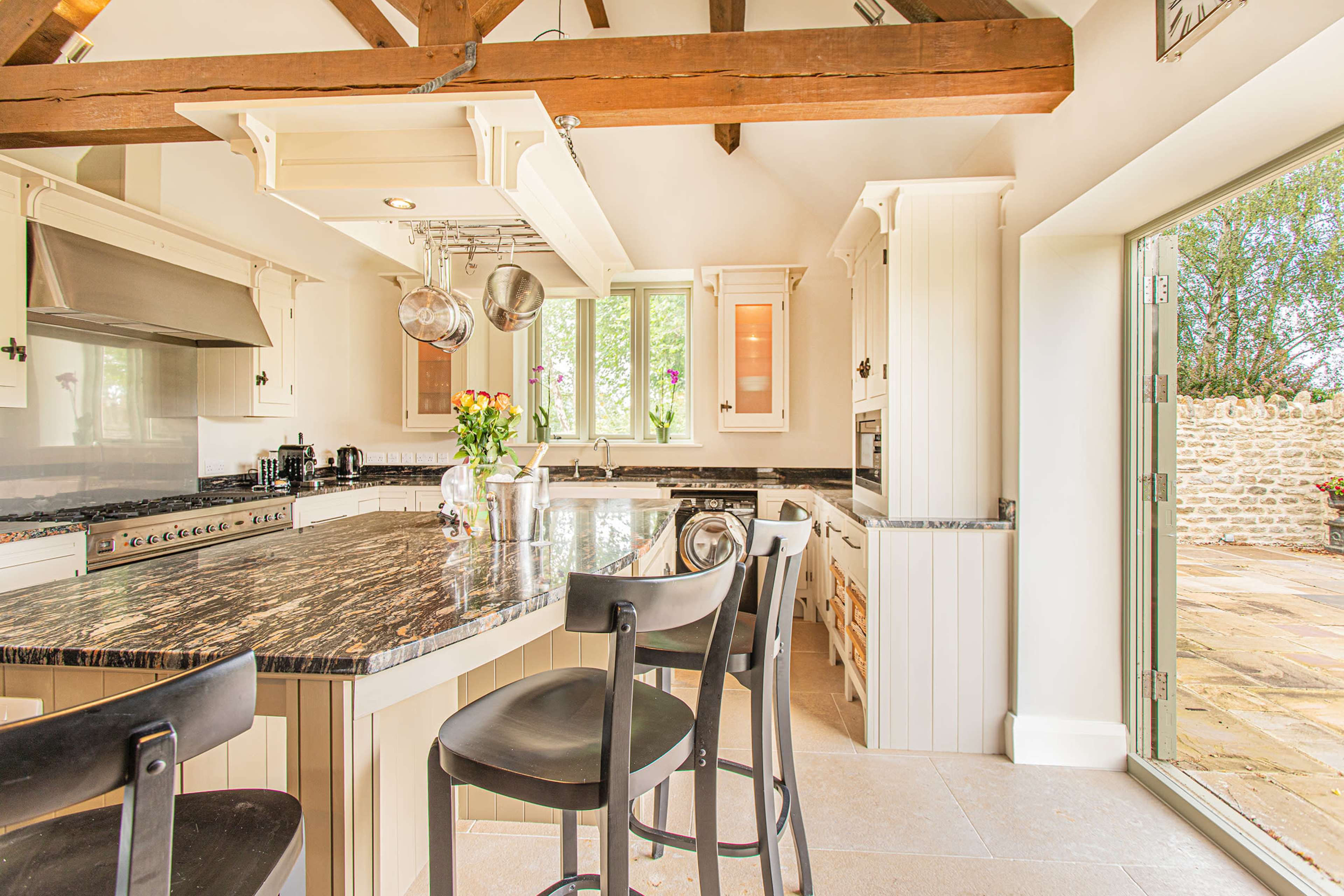 A modern kitchen features a large island with bar stools, stainless steel appliances, and opens to a stone patio through glass doors.