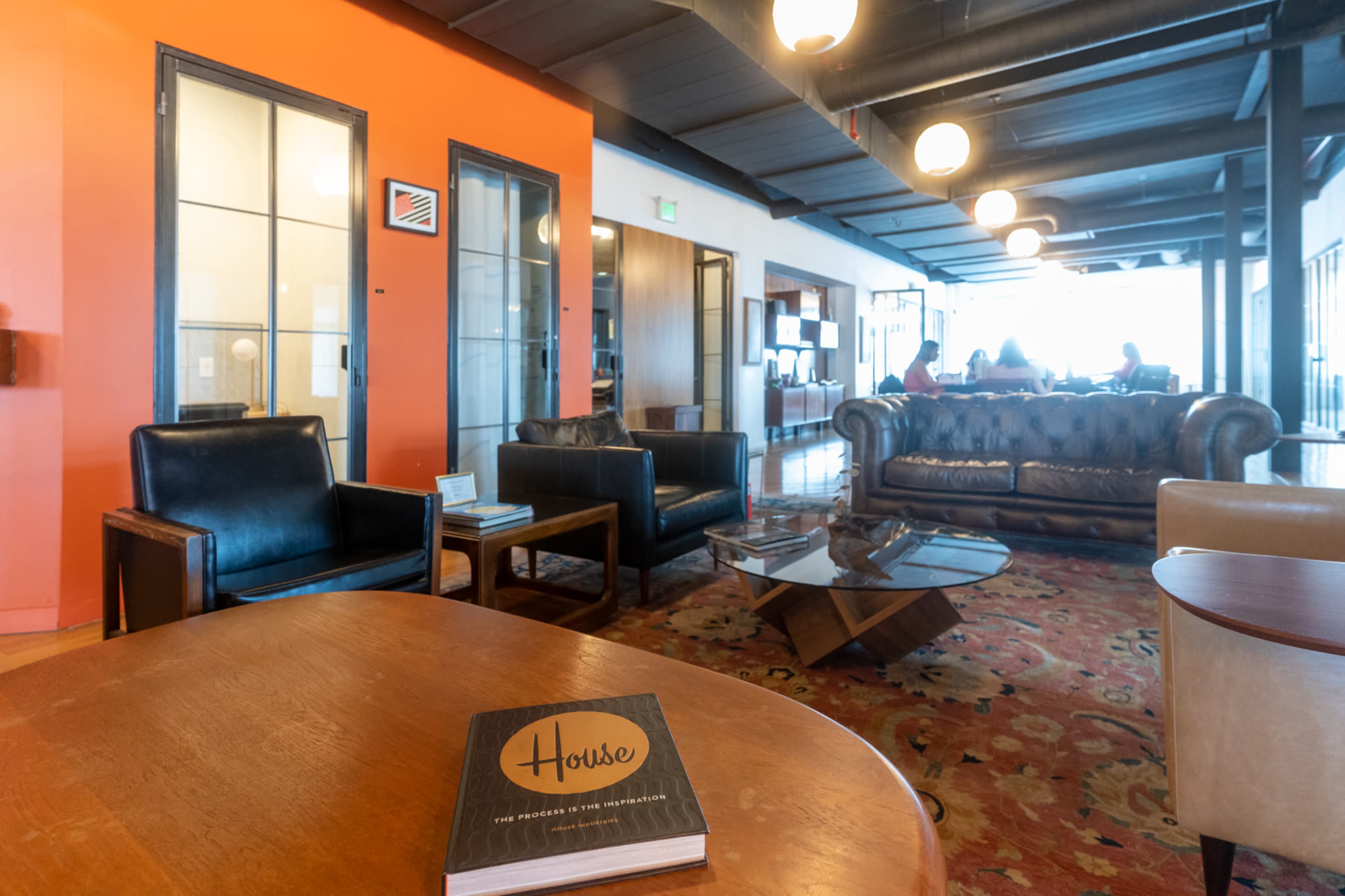 The image depicts a modern lounge area featuring leather armchairs, a wooden coffee table, and a patterned rug, with large windows in the background.