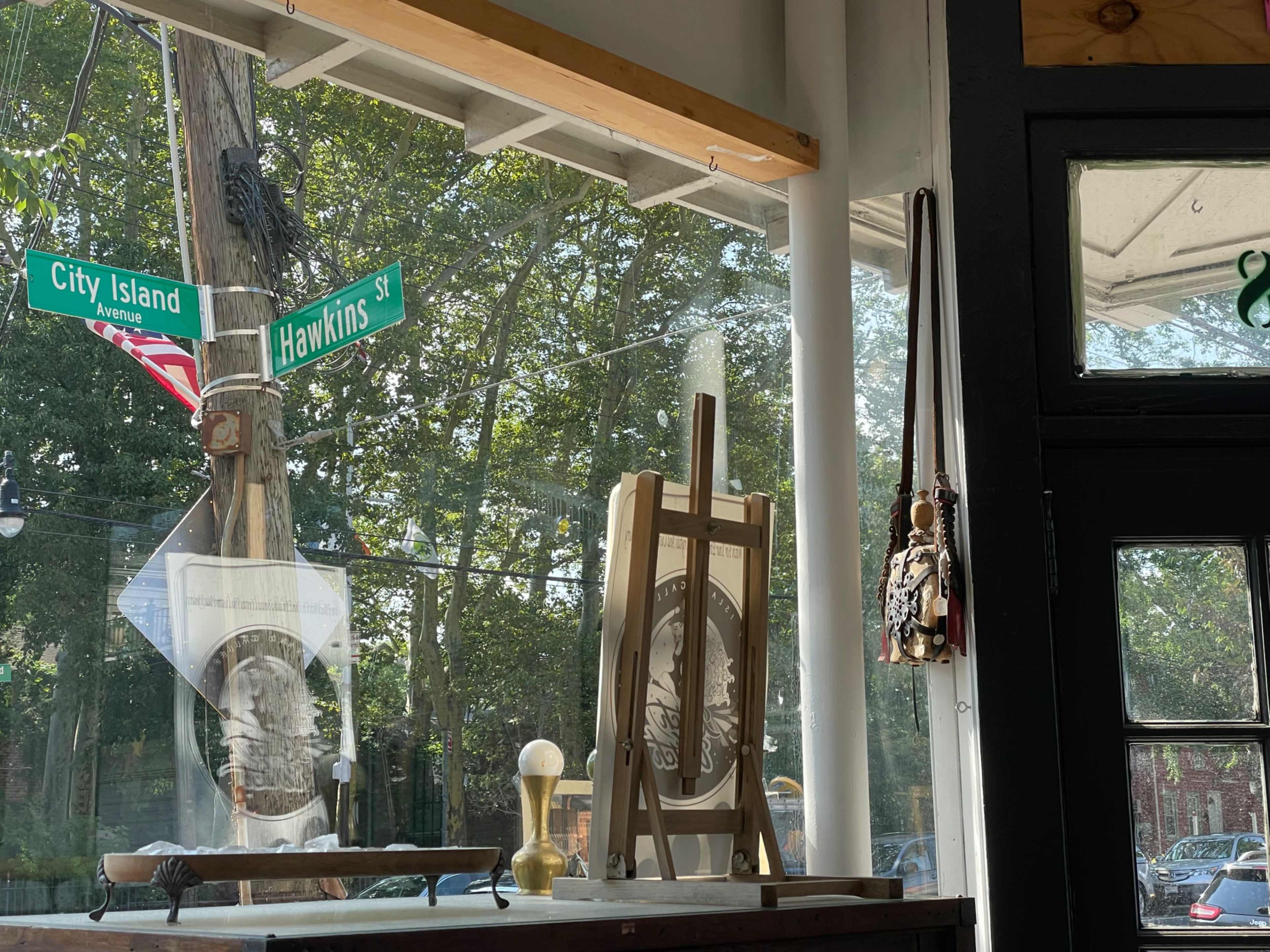 The image shows a view from inside a building looking out toward a street corner marked by City Island Avenue and Hawkins Street, with an artist's easel and decorative items visible in the foreground.