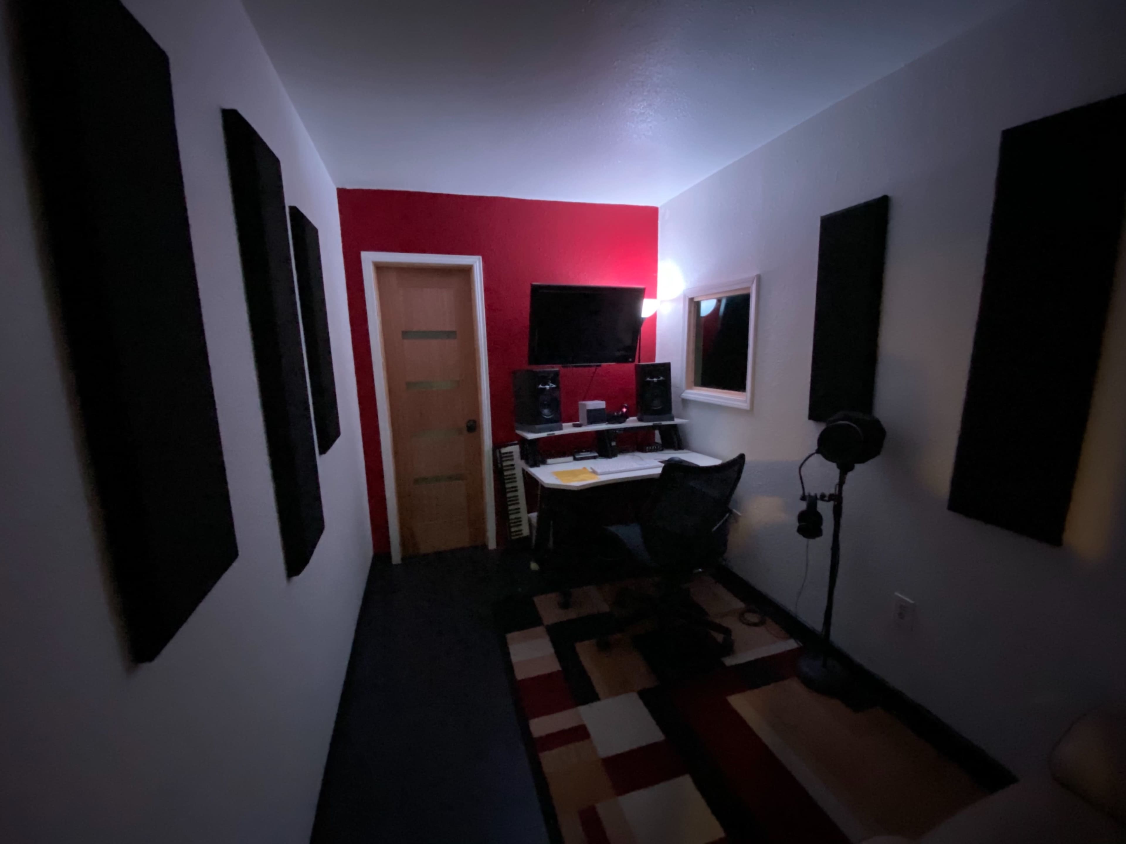 A dimly lit recording studio with soundproof panels on the walls, a desk with audio equipment, a monitor, and a microphone.