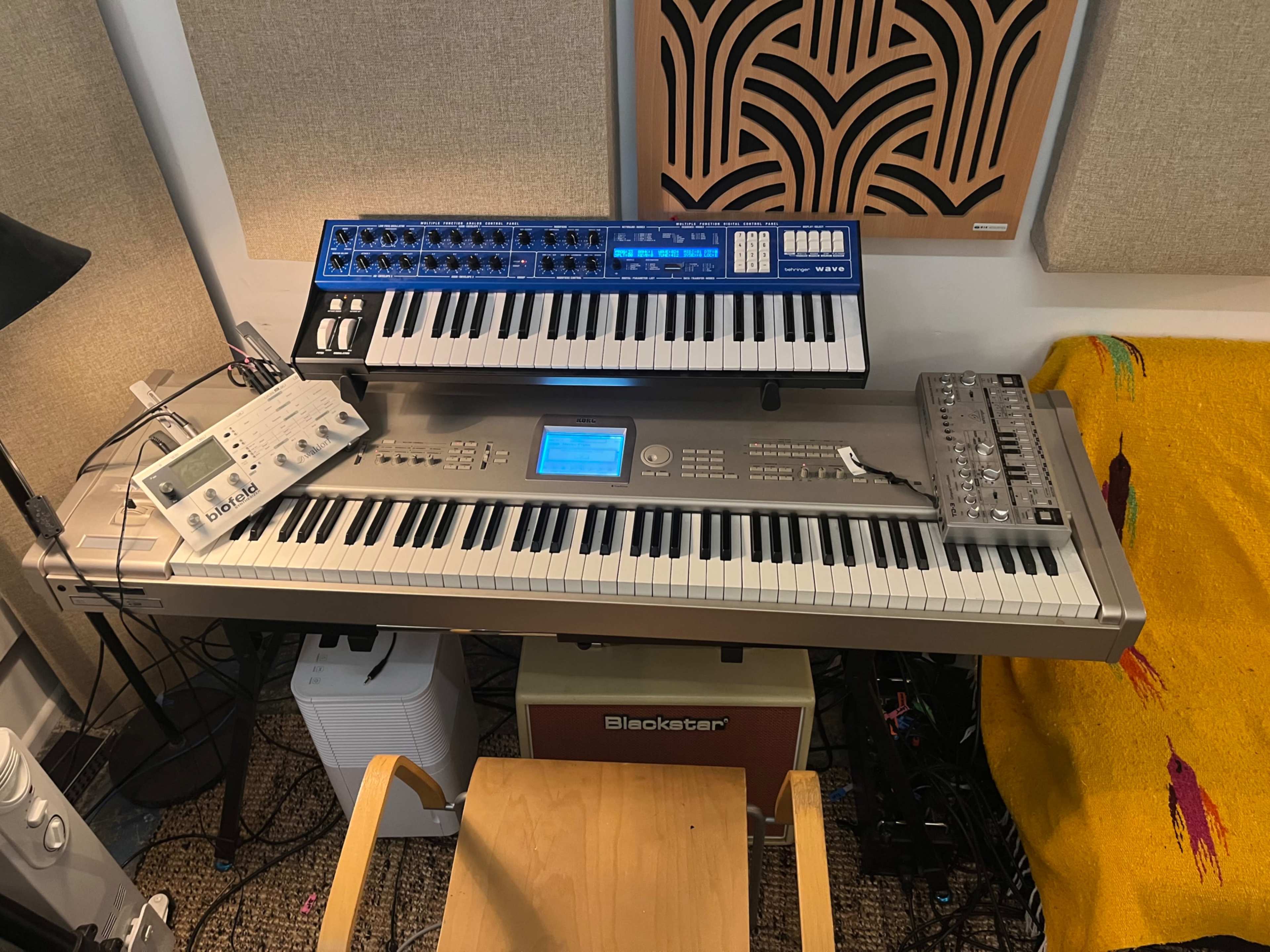 The image shows a music workstation featuring a keyboard, synthesizers, and a drum machine arranged on a table in a studio setting.