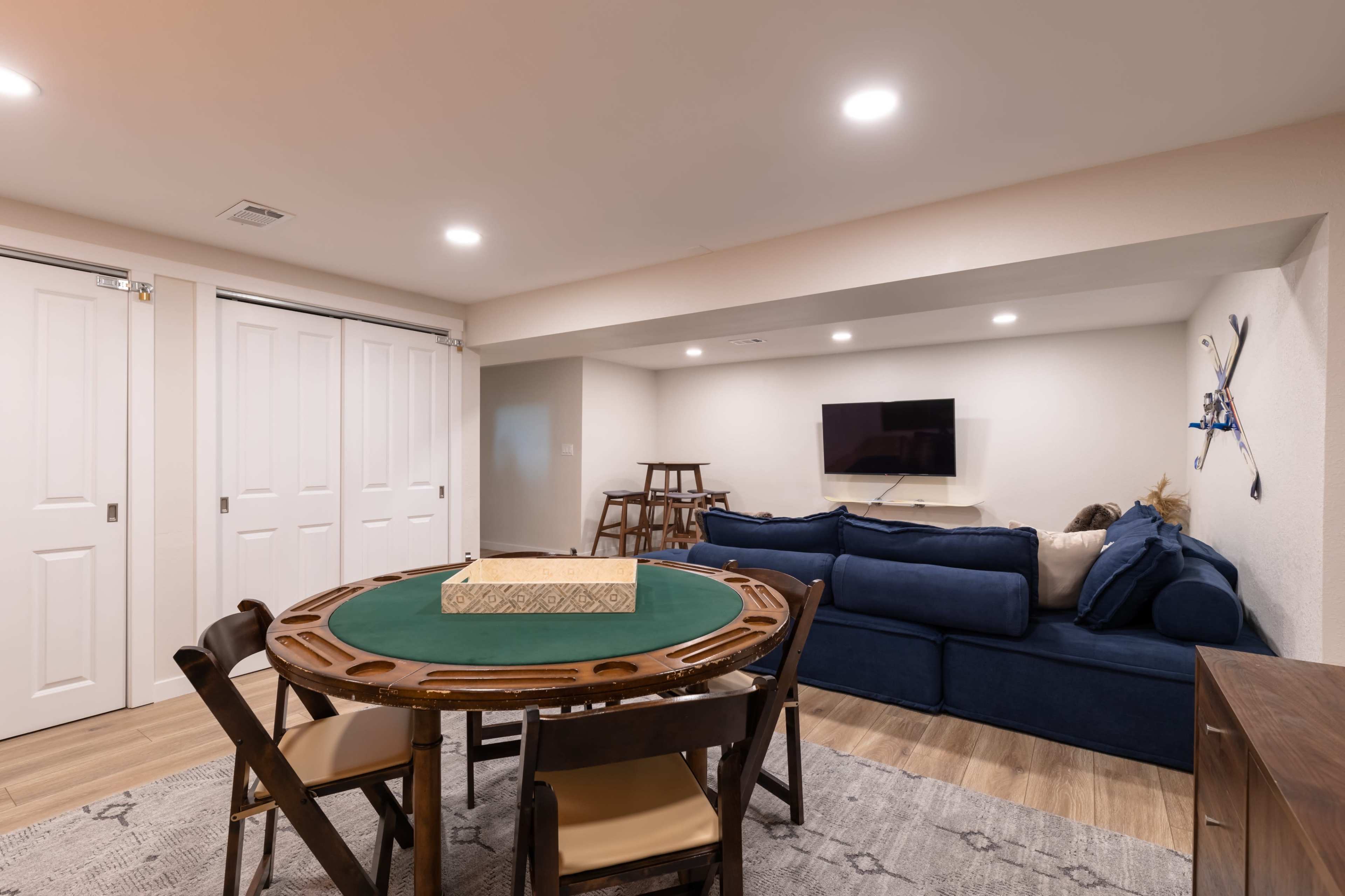 A room with a round poker table at the center, surrounded by chairs, and a blue sofa against the wall, with a television mounted above it.