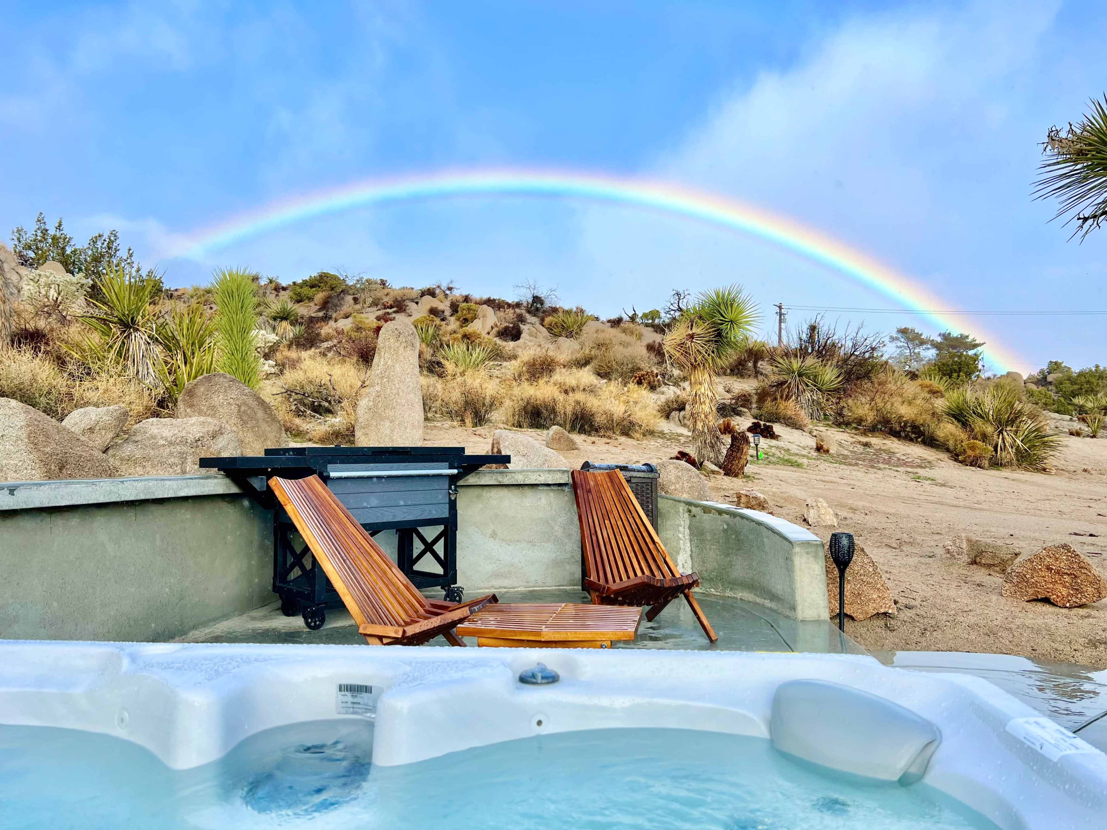 A hot tub with two wooden chairs overlooks a desert landscape with a rainbow arching in the sky.