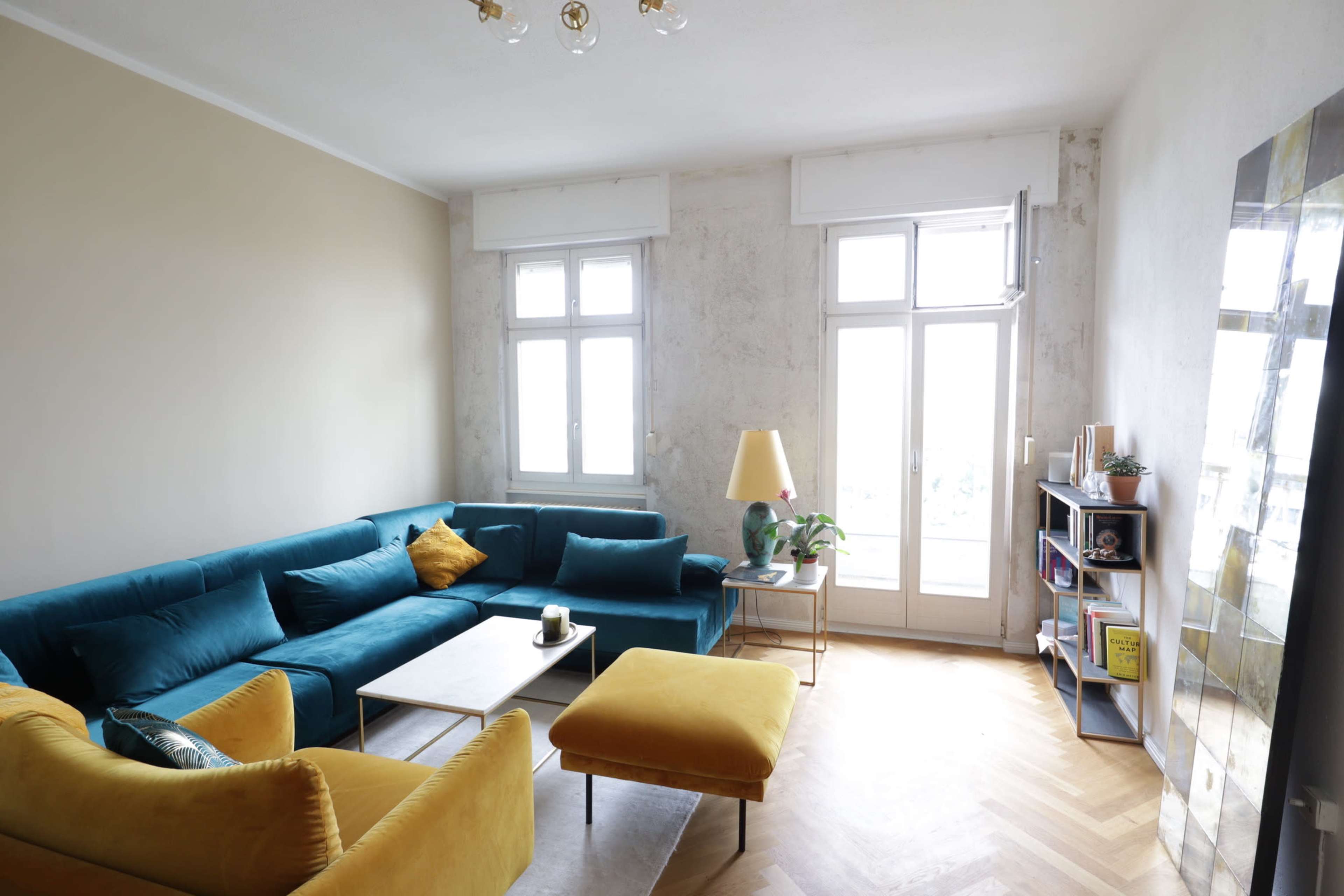 The living room features a mix of teal and yellow furniture, large windows allowing natural light, and a bookshelf against a textured wall.
