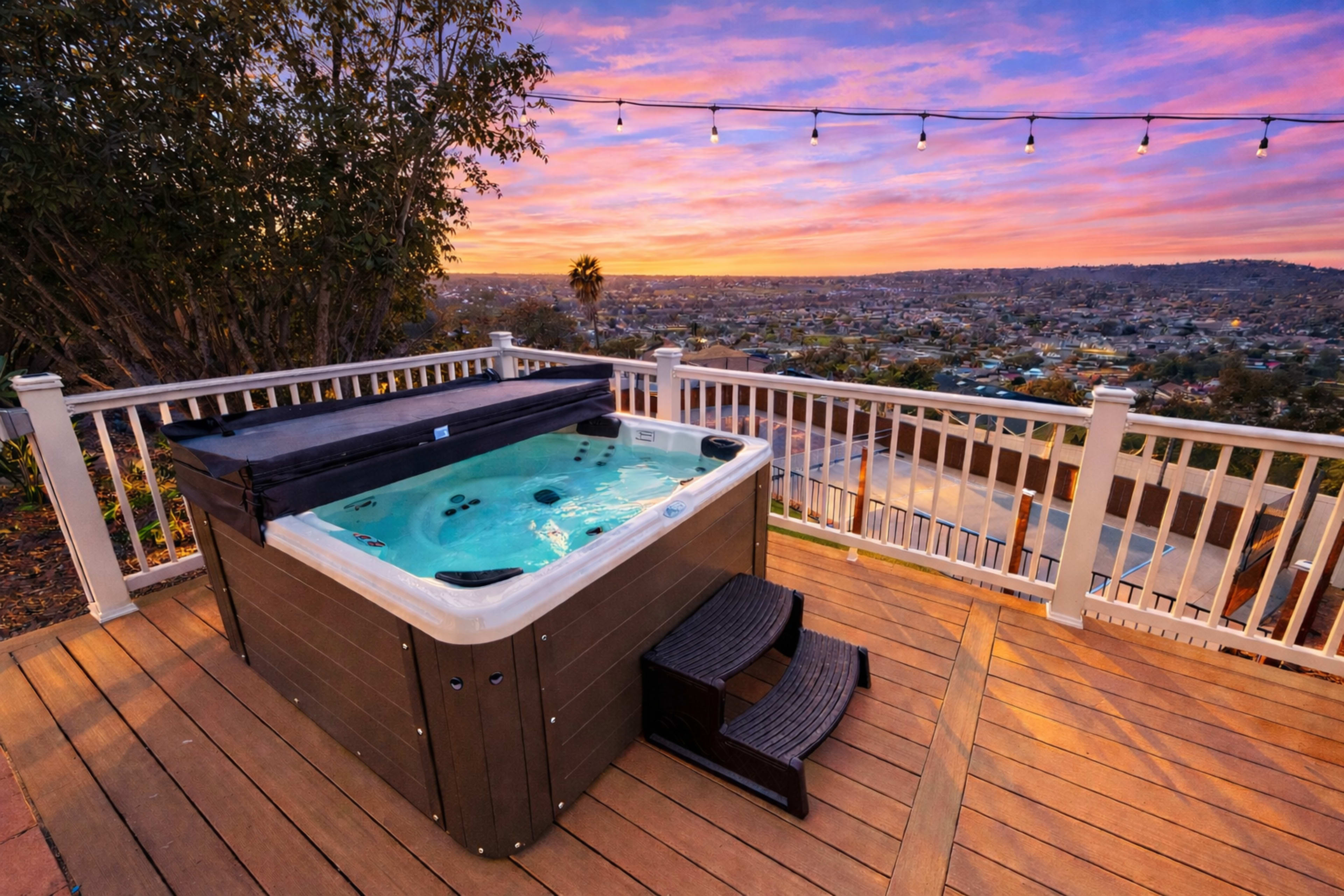 A hot tub sits on a wooden deck overlooking a cityscape during sunset, with string lights illuminating the area.