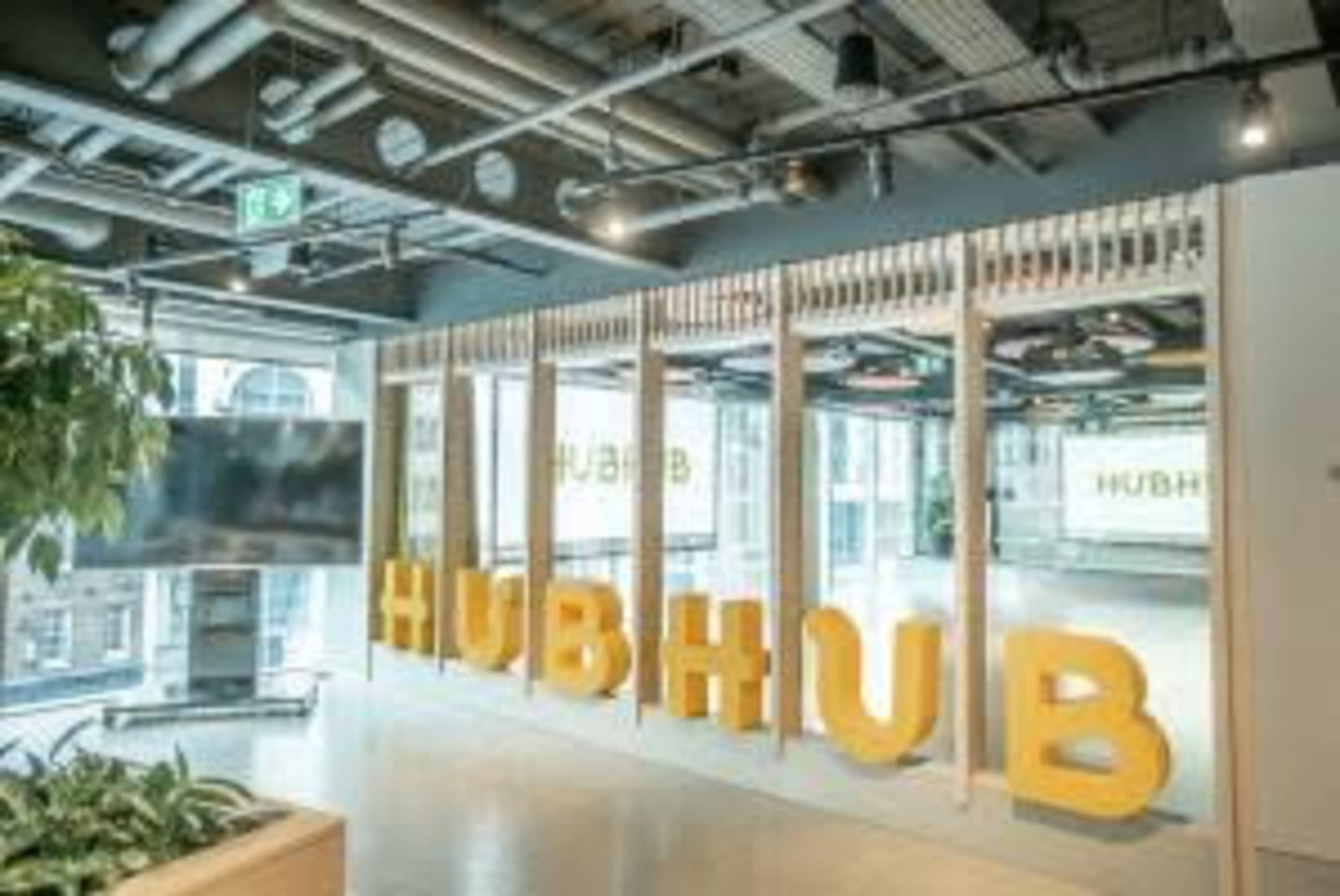 The image shows a modern office space with large yellow letters spelling "HUBHUB" and a television screen in the background.