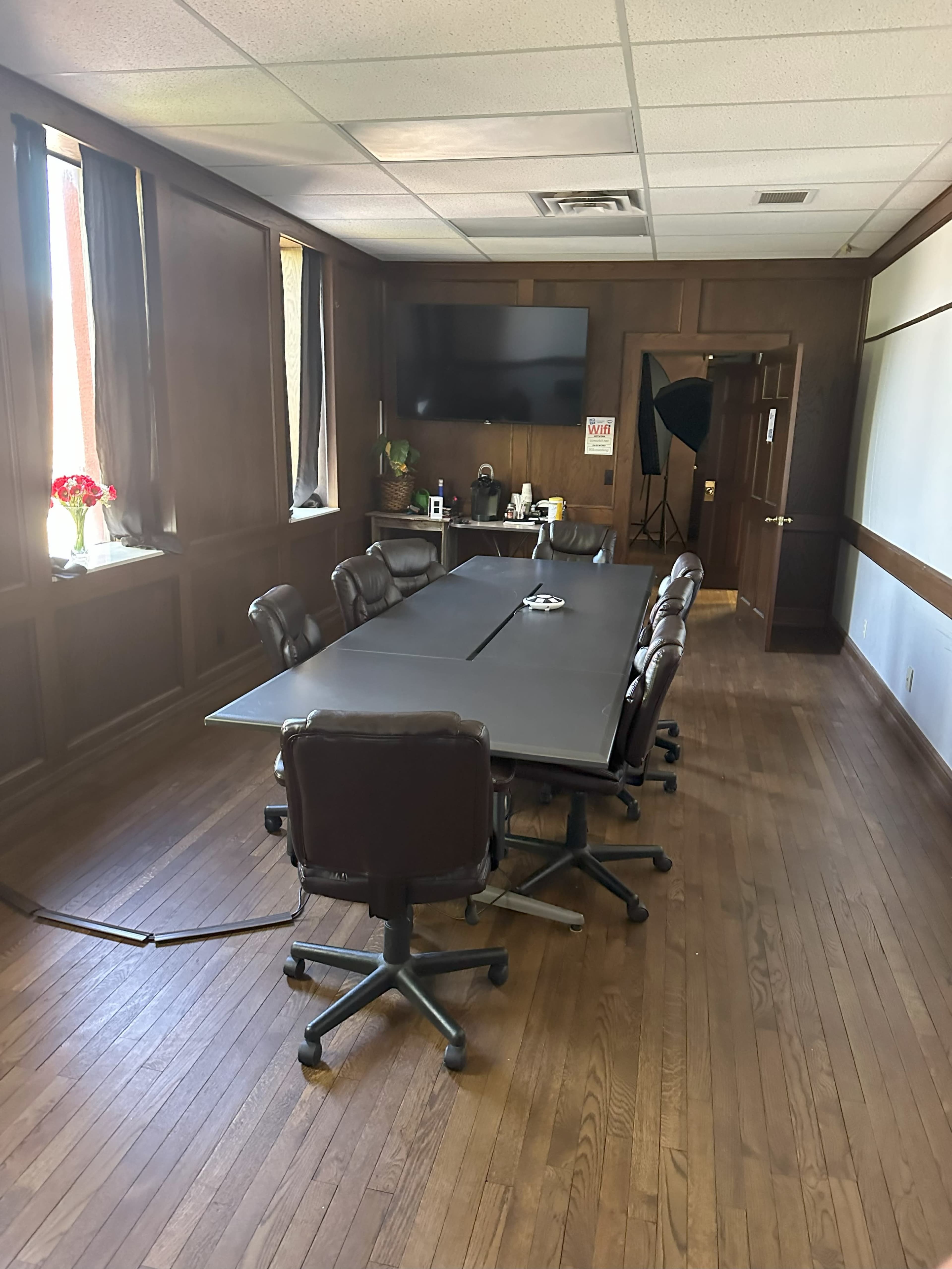 A conference room features a long table with rolling chairs, wooden paneling, and a large screen on the wall.