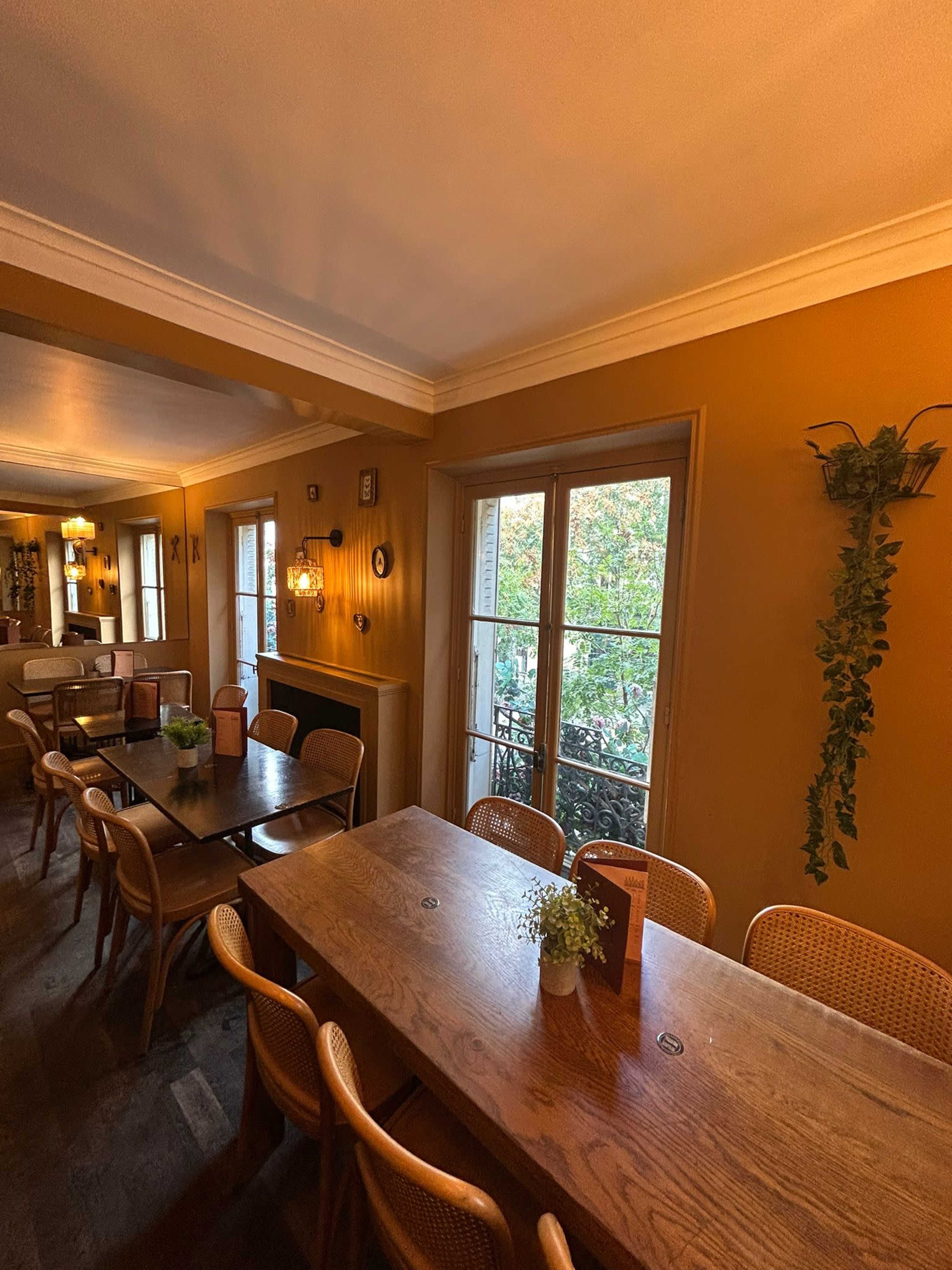 The image shows a cozy dining area with wooden tables and chairs, large windows opening to a view of greenery outside.