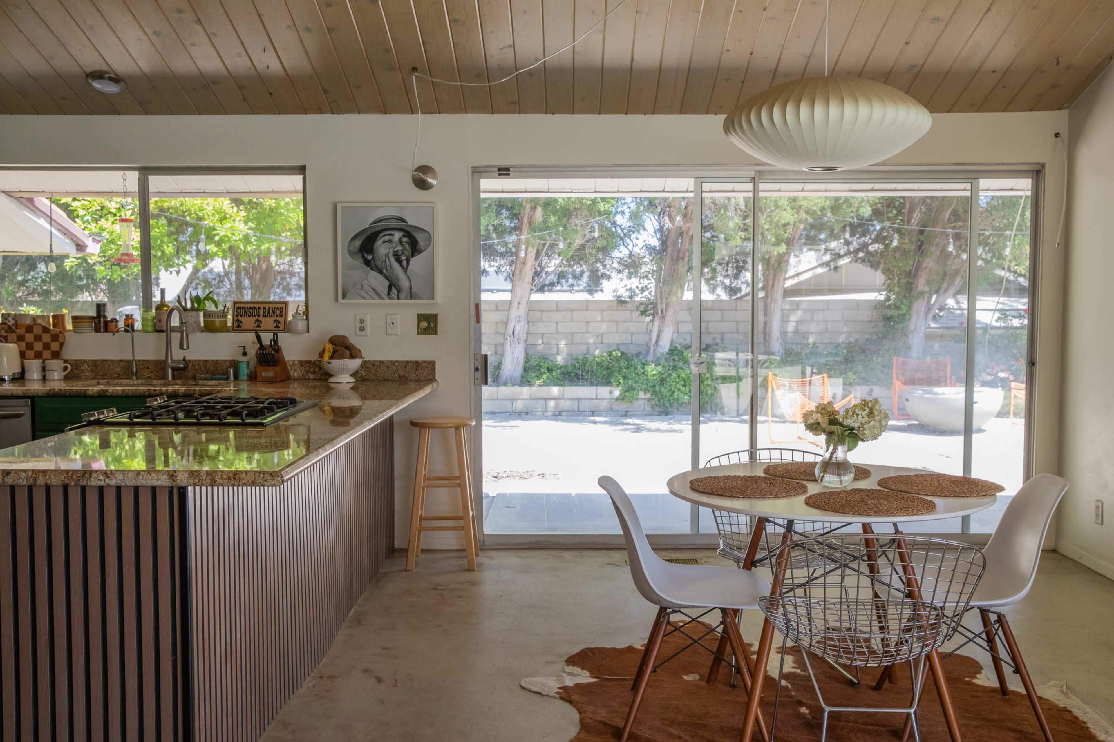 A modern kitchen and dining area features a round table with white chairs, a large window overlooking a patio, and a countertop with bar stools.