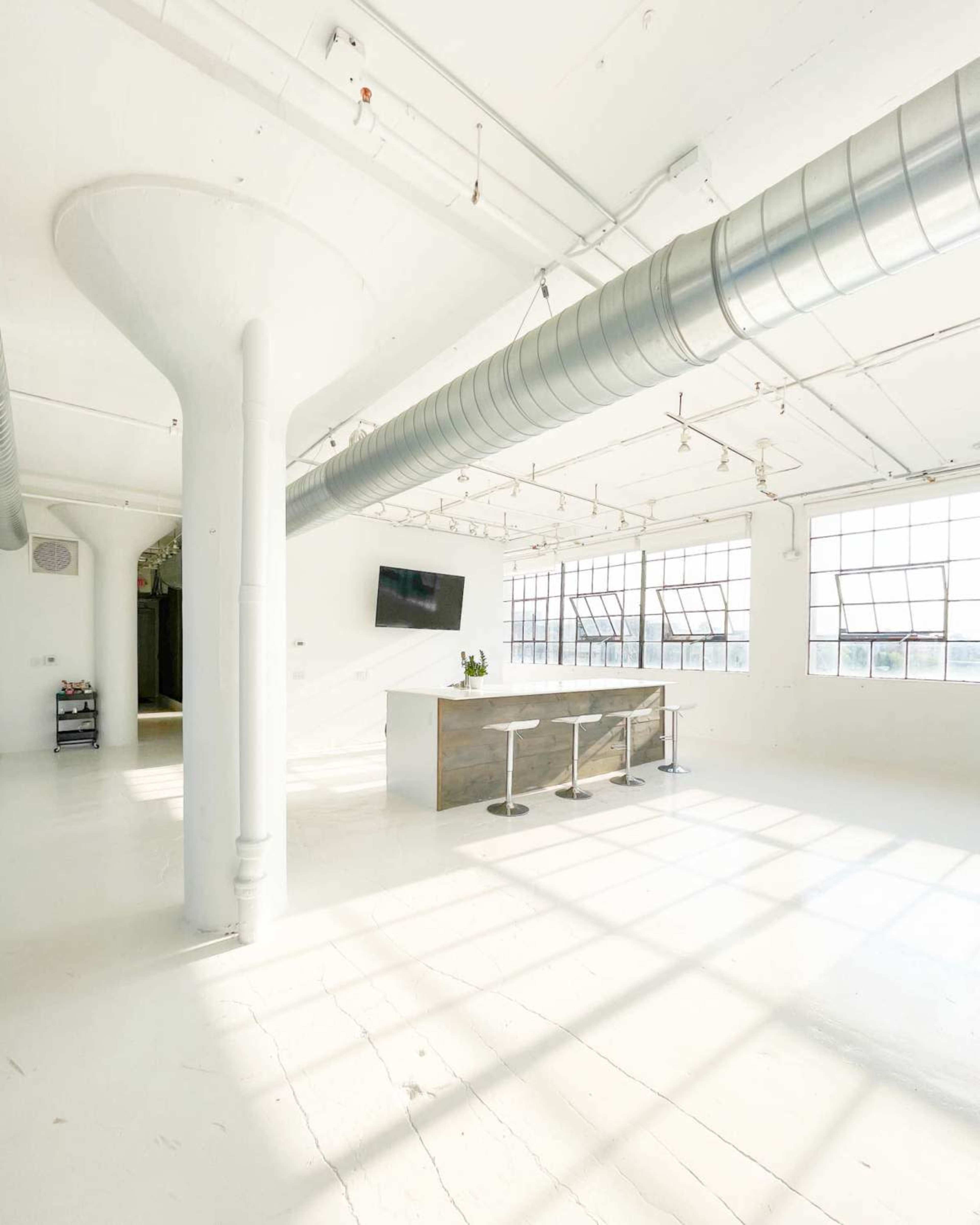 The image shows a spacious, well-lit industrial-style room with white walls and a central bar area featuring three stools.
