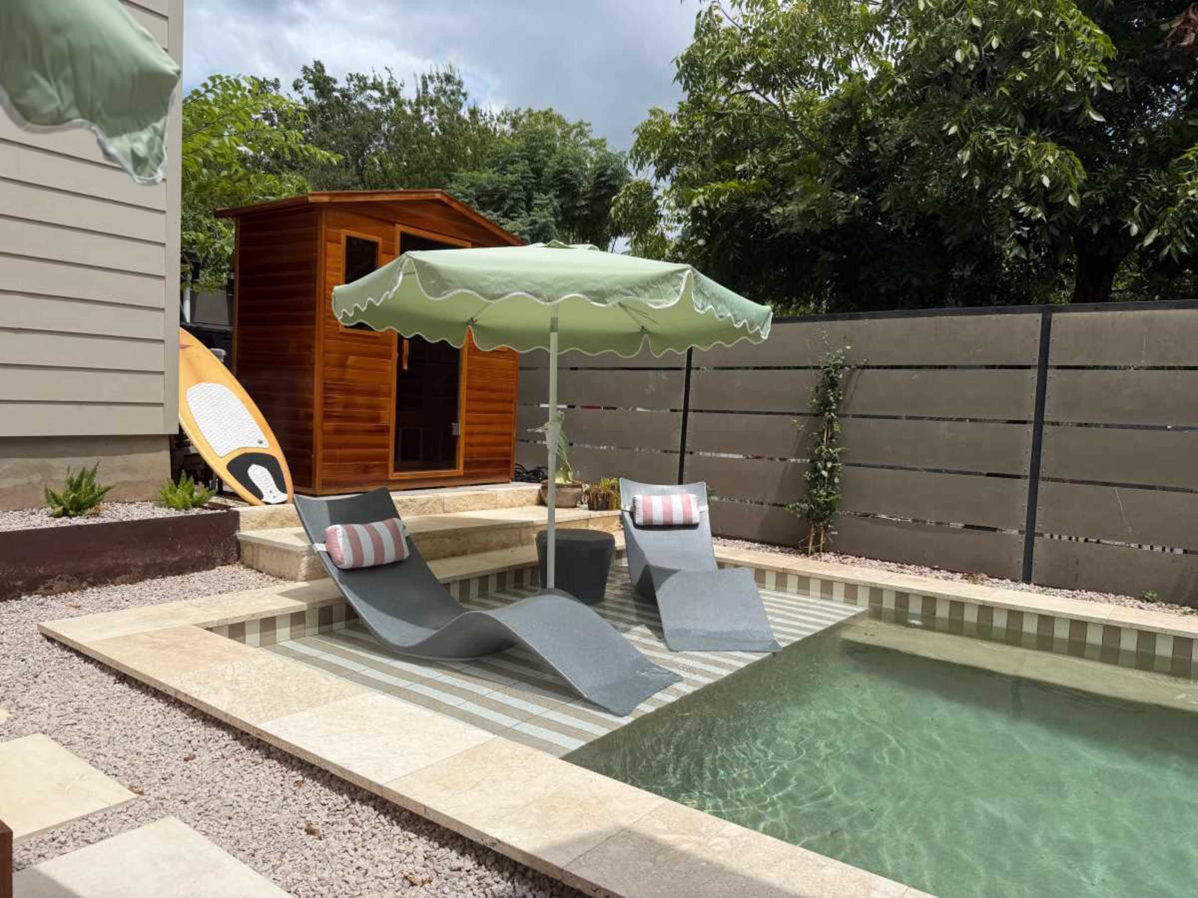 A small swimming pool is surrounded by two lounge chairs and an umbrella, with a wooden shed and a paddleboard in the background.