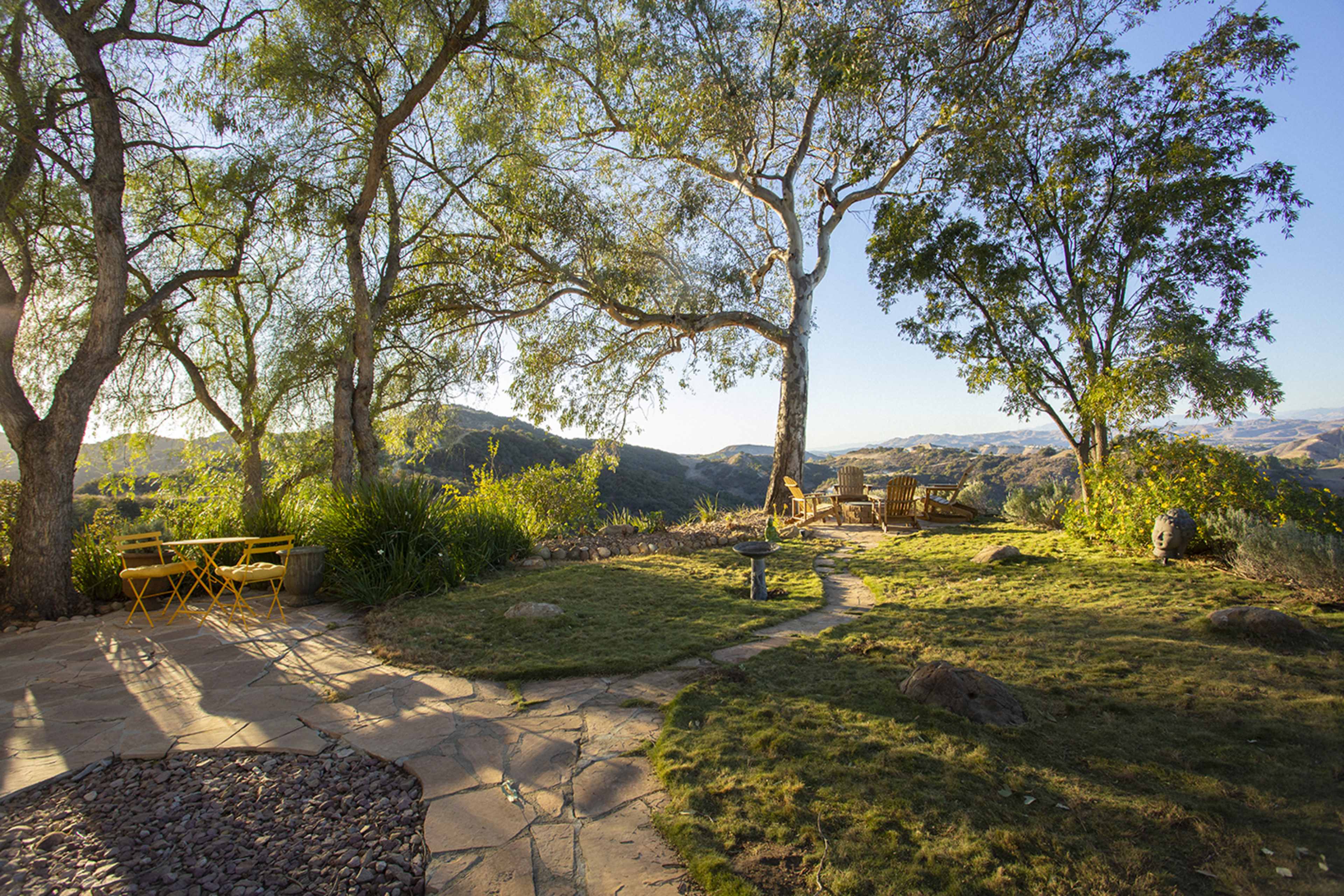 A stone path curves through a grassy area surrounded by trees, leading to seating and a table with a scenic view of distant hills.