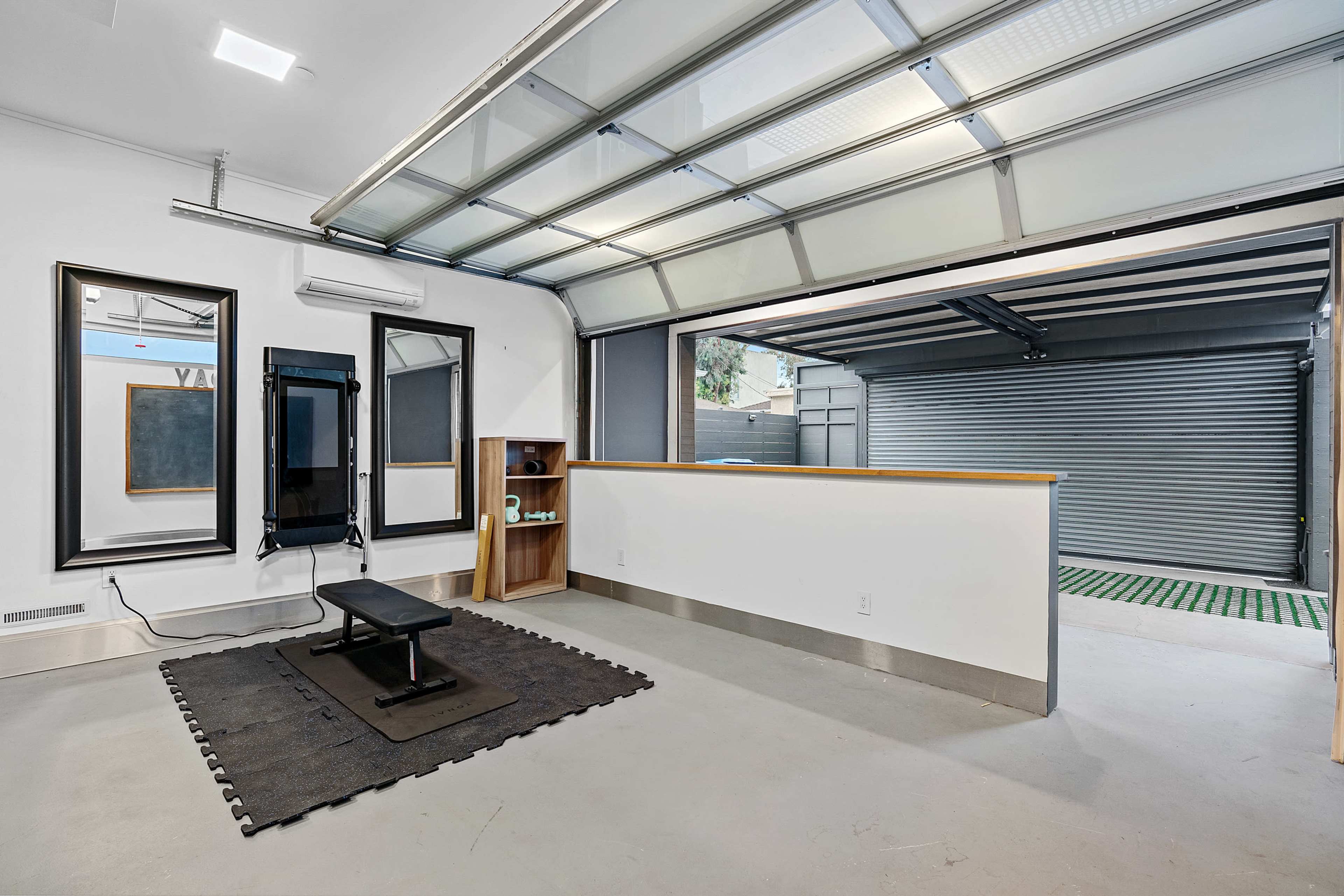 The image shows a spacious garage with large glass doors, minimalistic furniture, and a simple layout, including a worktable and a mat on the floor.