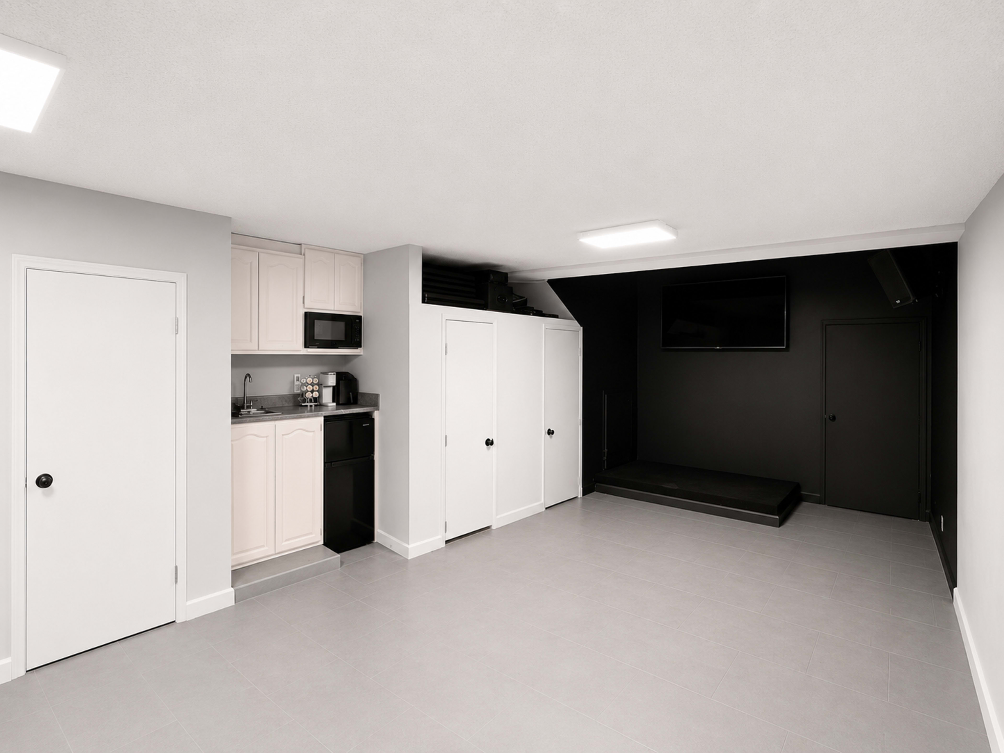 The image shows a modern, minimally furnished basement with a kitchenette on one side and a small stage area against a black wall.