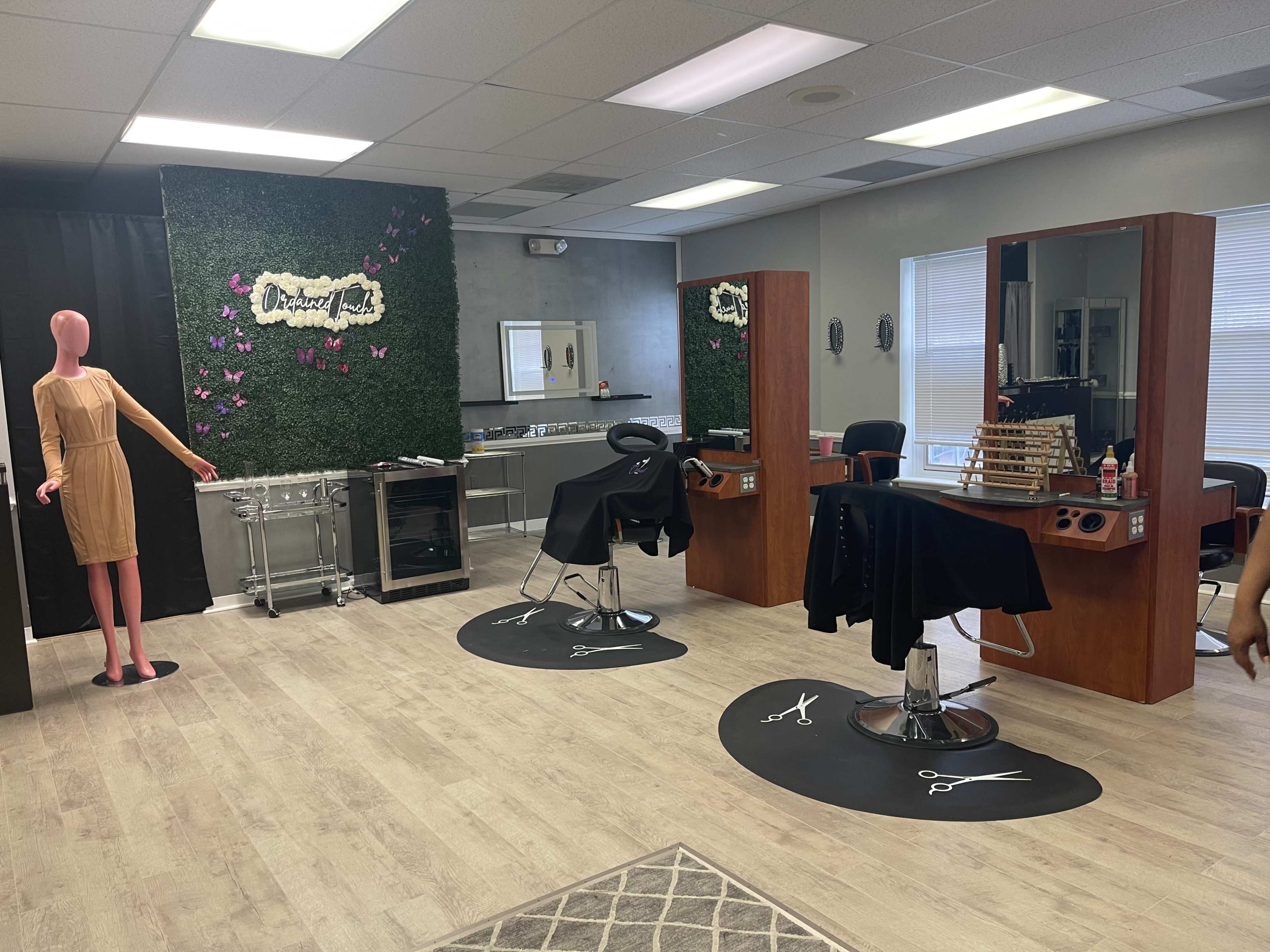 A hair salon with two styling stations, a green wall adorned with decorations, and a mannequin dressed in a brown outfit.