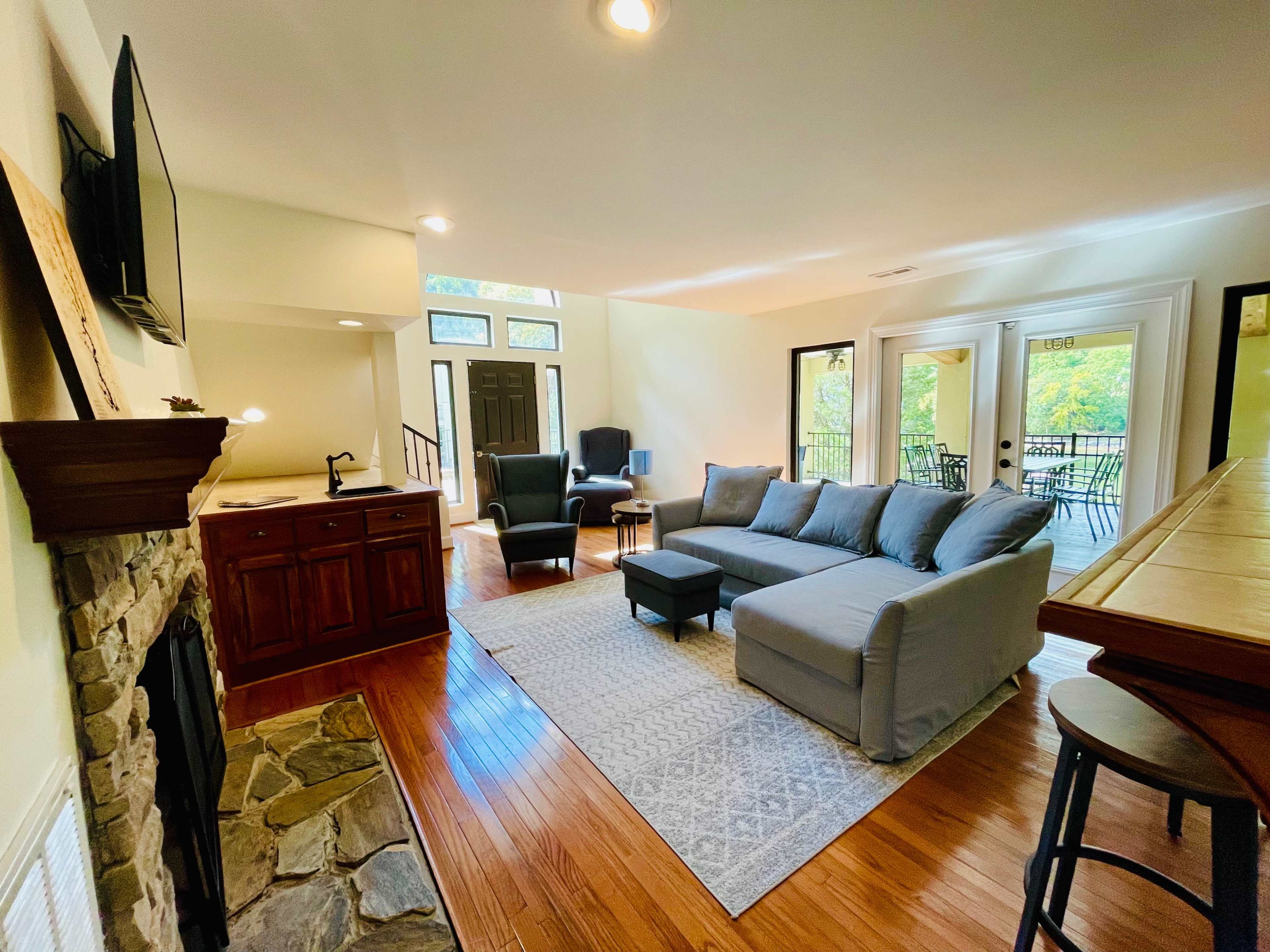 The image shows a living room with a gray sectional sofa, a stone fireplace, and a view of large windows leading to an outdoor patio.