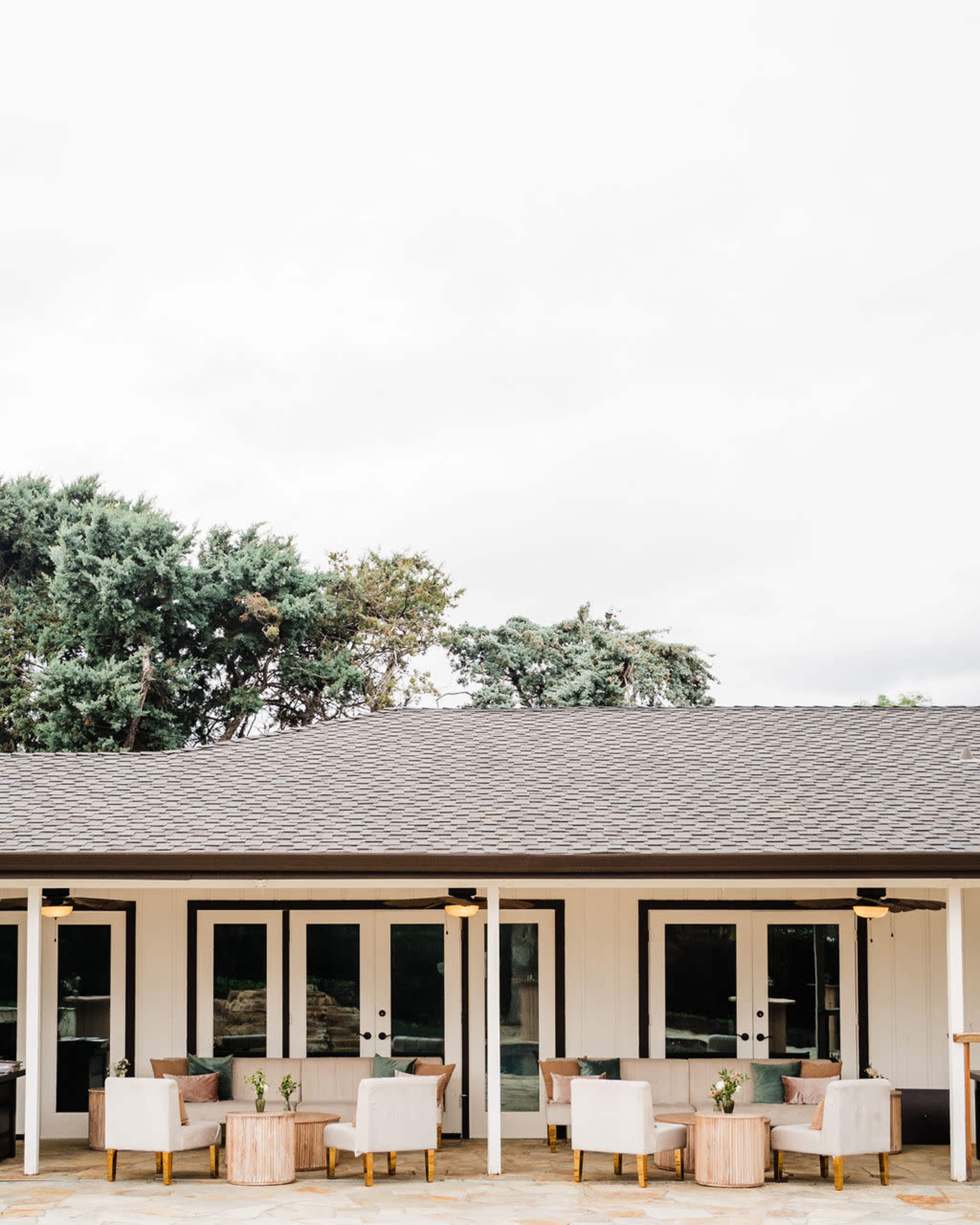 A modern outdoor seating area features four white chairs with gold accents arranged around small wooden tables in front of a building with a gray roof and large glass doors.