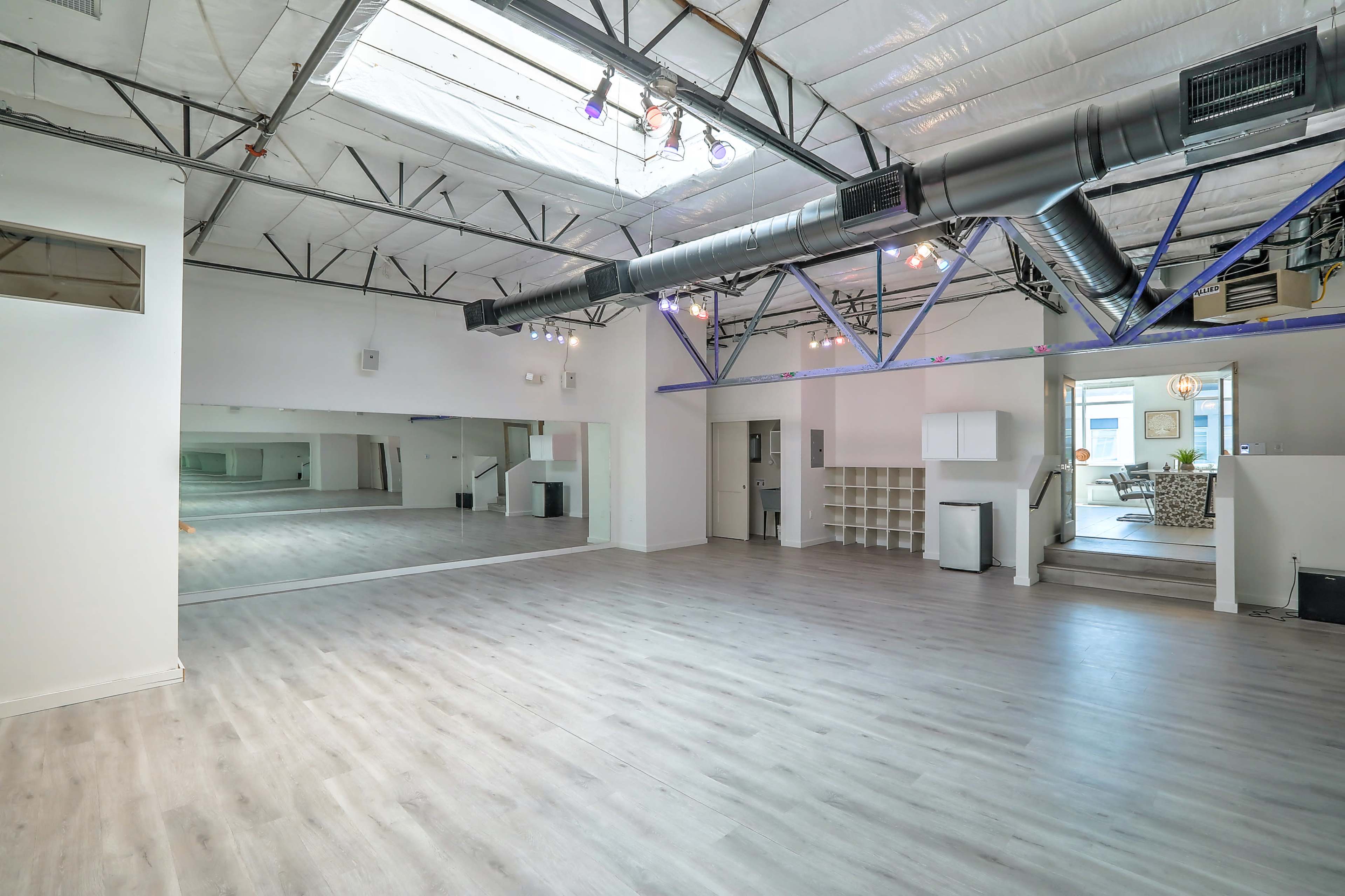 A spacious, well-lit room with a mirrored wall, wooden flooring, and exposed ductwork on the ceiling.