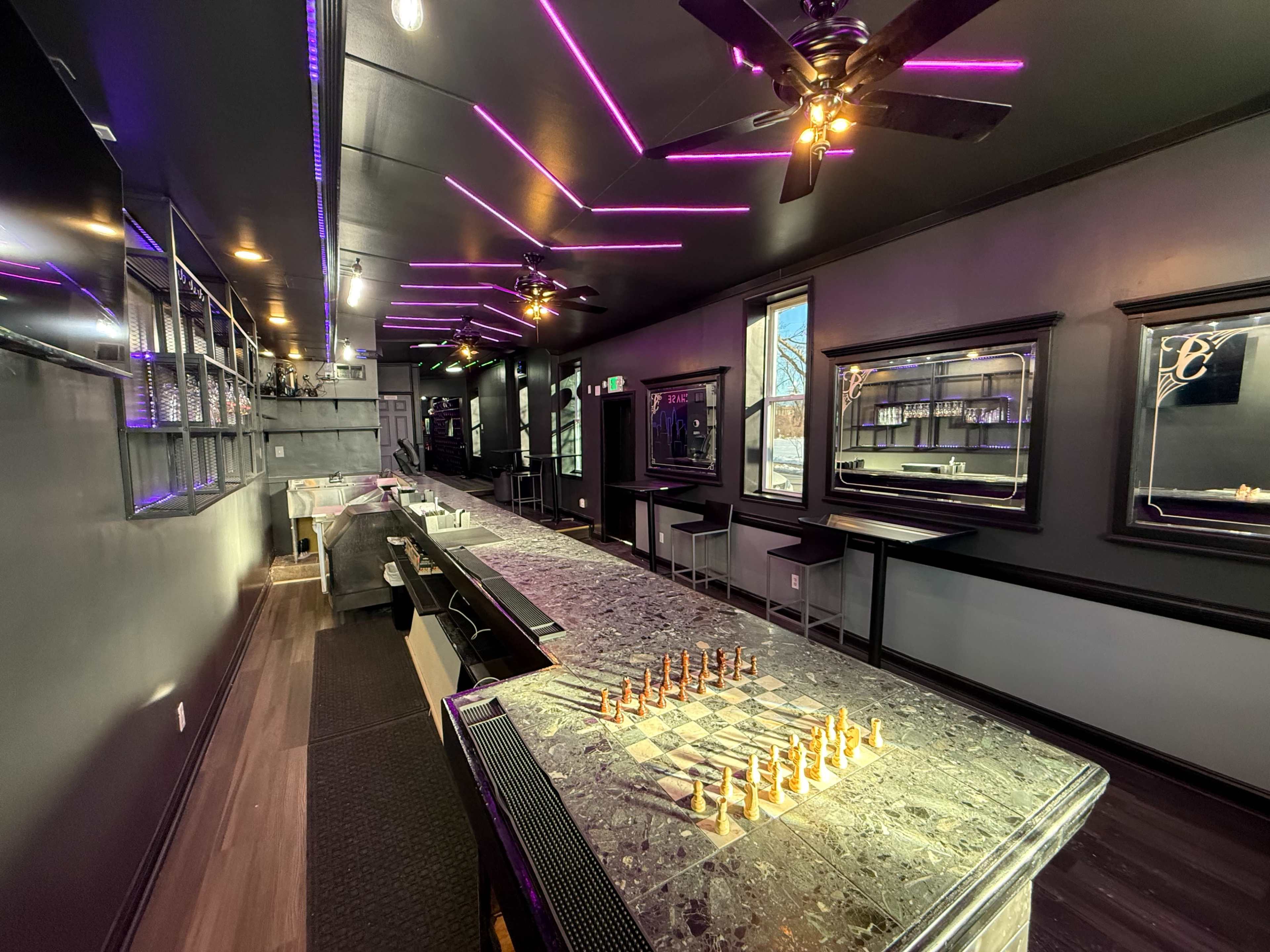 The image shows a modern bar with a sleek, dark interior featuring purple LED lighting and a chess set on the countertop.