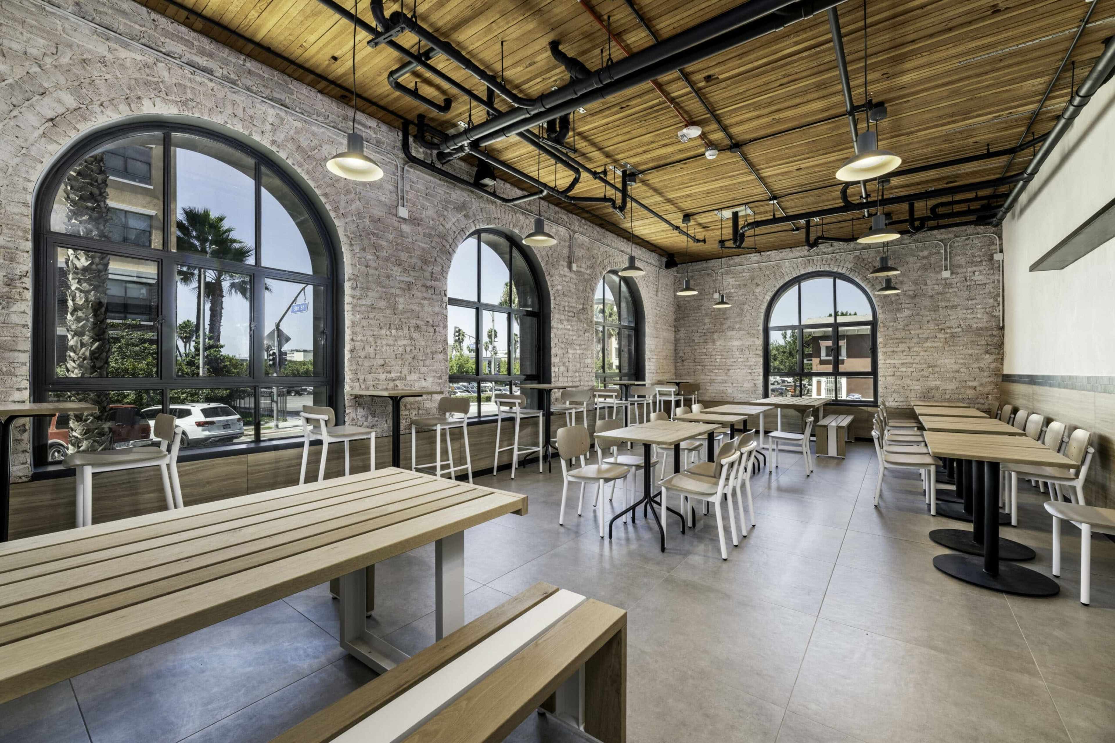 A spacious, modern café interior with large arched windows, wooden ceilings, and a mix of tables and chairs arranged neatly.