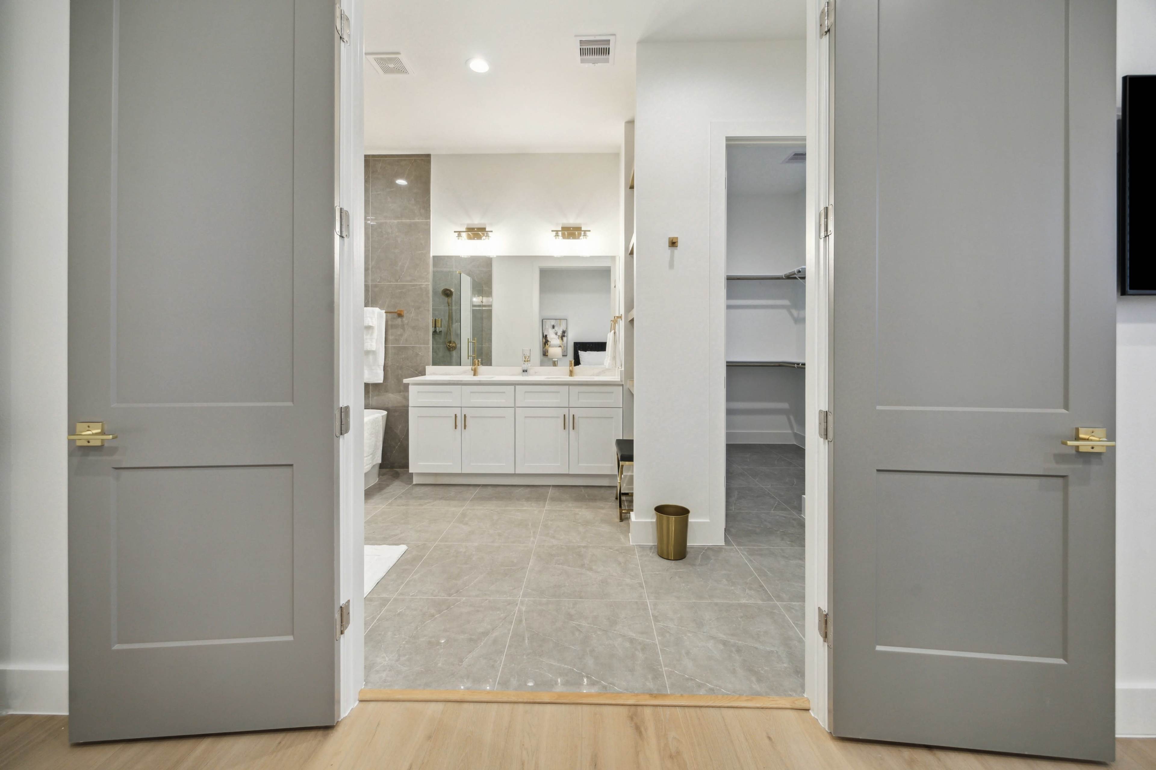 A spacious, modern bathroom is visible through two open gray doors, featuring a double vanity, shower area, and adjacent walk-in closet.