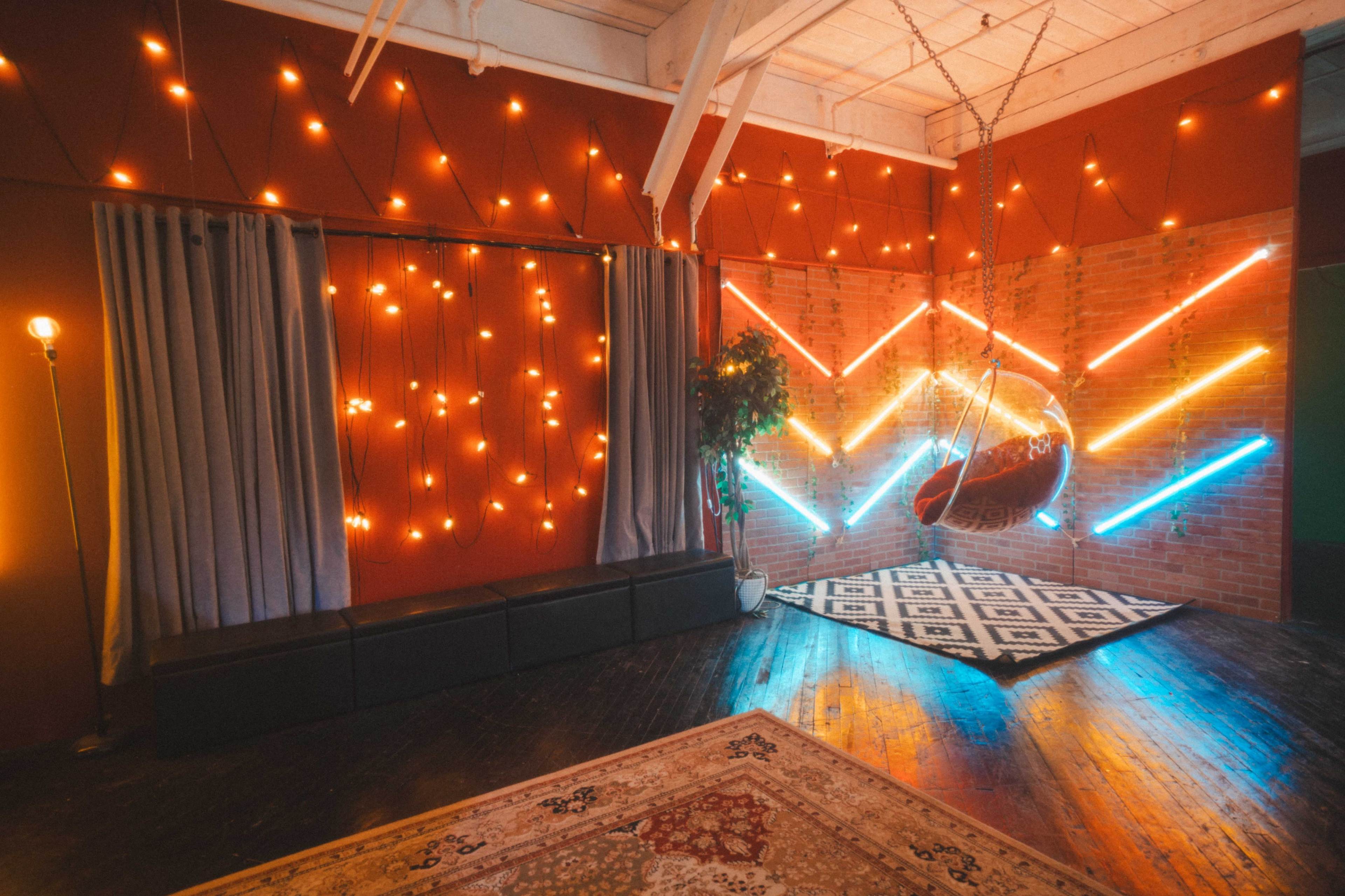 The image shows a cozy room with warm string lights, patterned rugs, a swing chair, and a minimalist black sofa against a brightly lit wall.