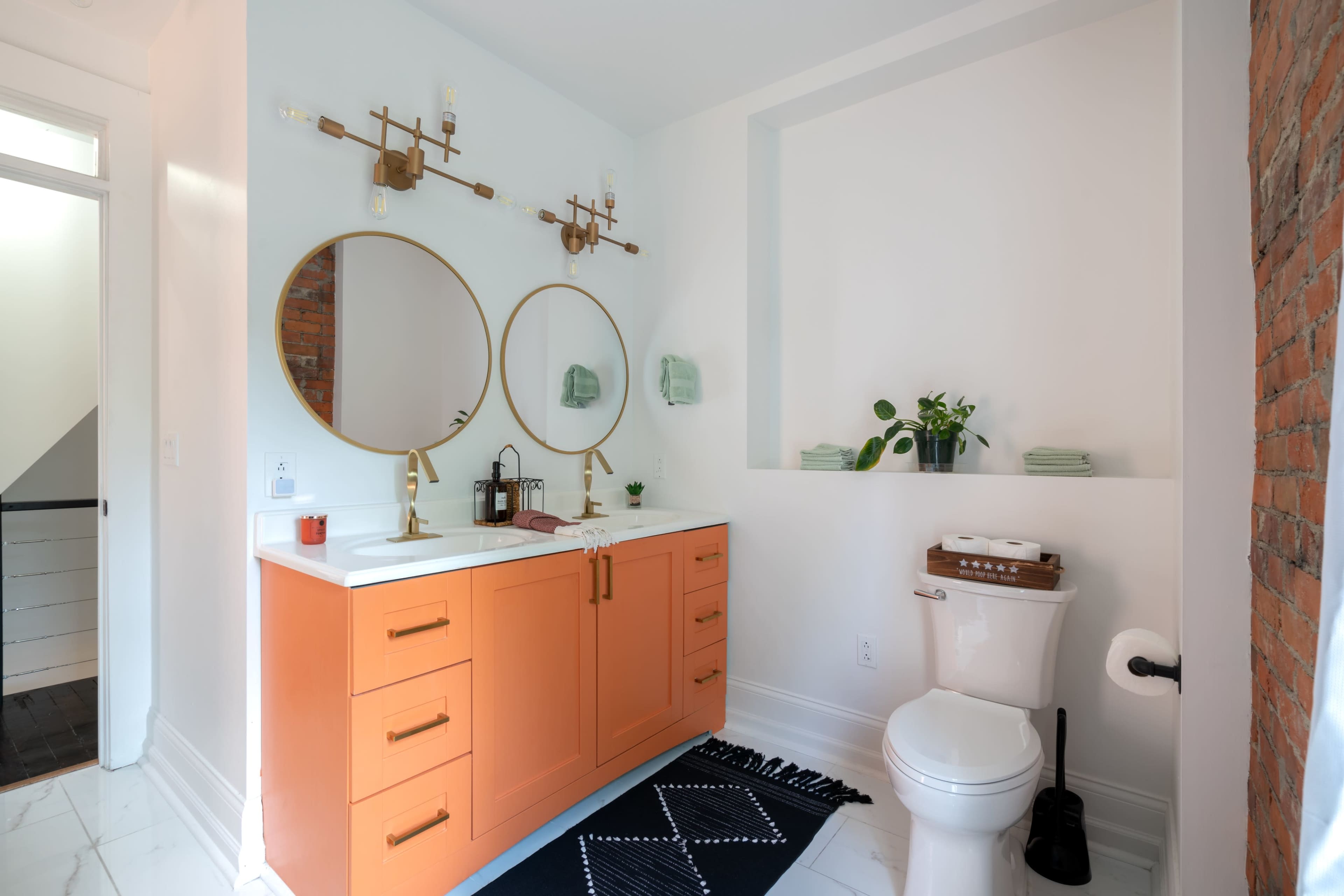 A modern bathroom featuring two round mirrors above a double sink with orange cabinetry, a toilet, and decorative plants against a white and brick wall.