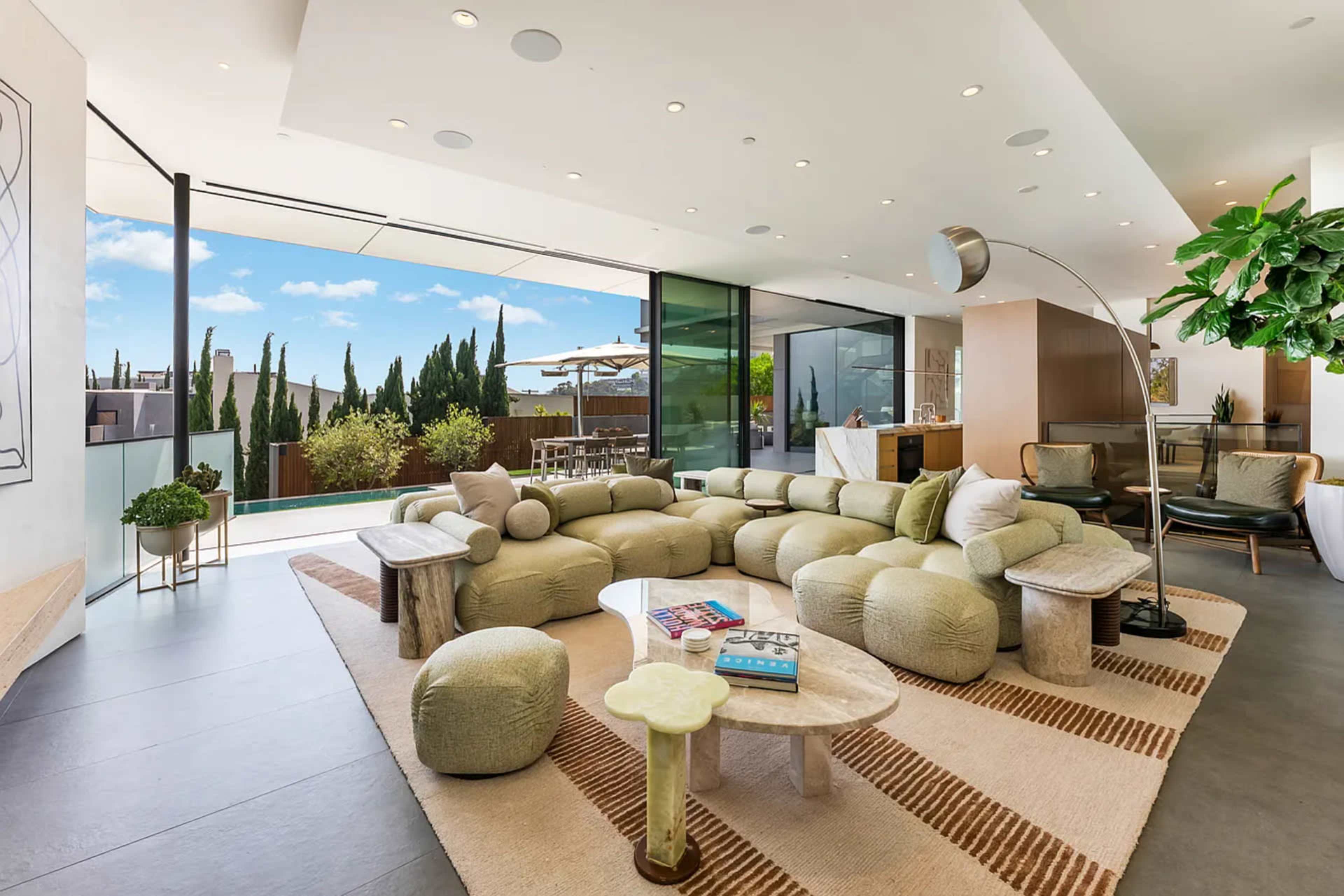 The image shows a modern living room with a unique, rounded sectional sofa, a coffee table, and large windows that open to a patio area with greenery.