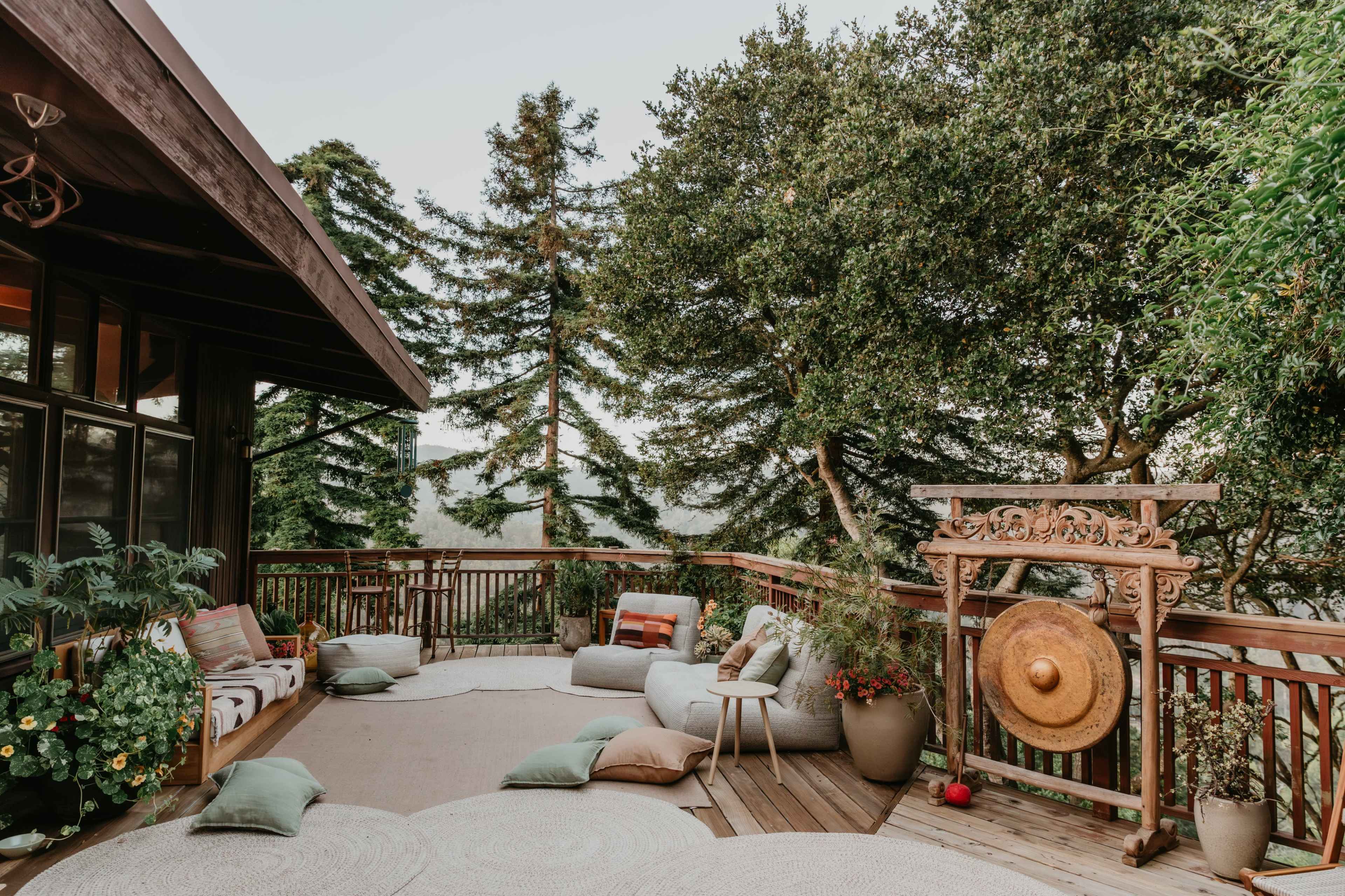 A wooden deck features various seating areas with cushions, plants, and a large decorative gong against a backdrop of tall trees.