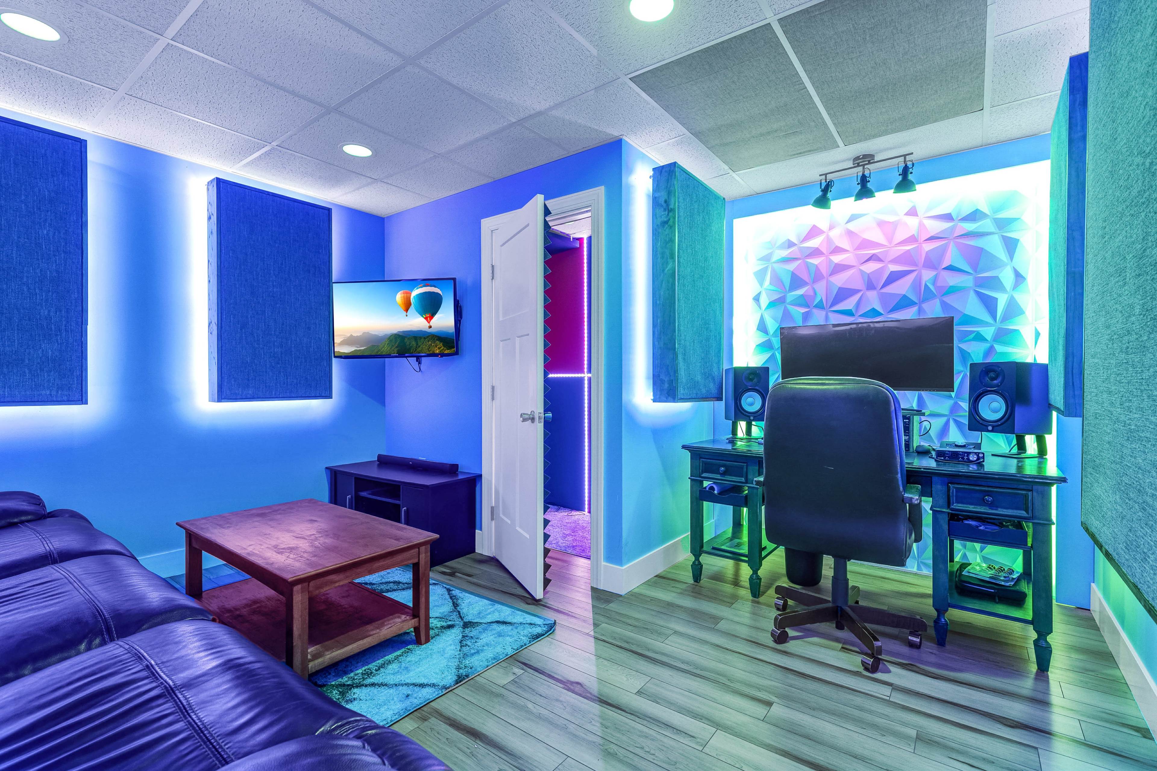 A modern room with a gaming setup, featuring a desk with monitors, a black ergonomic chair, blue walls, and acoustic panels, along with a couch and a television.