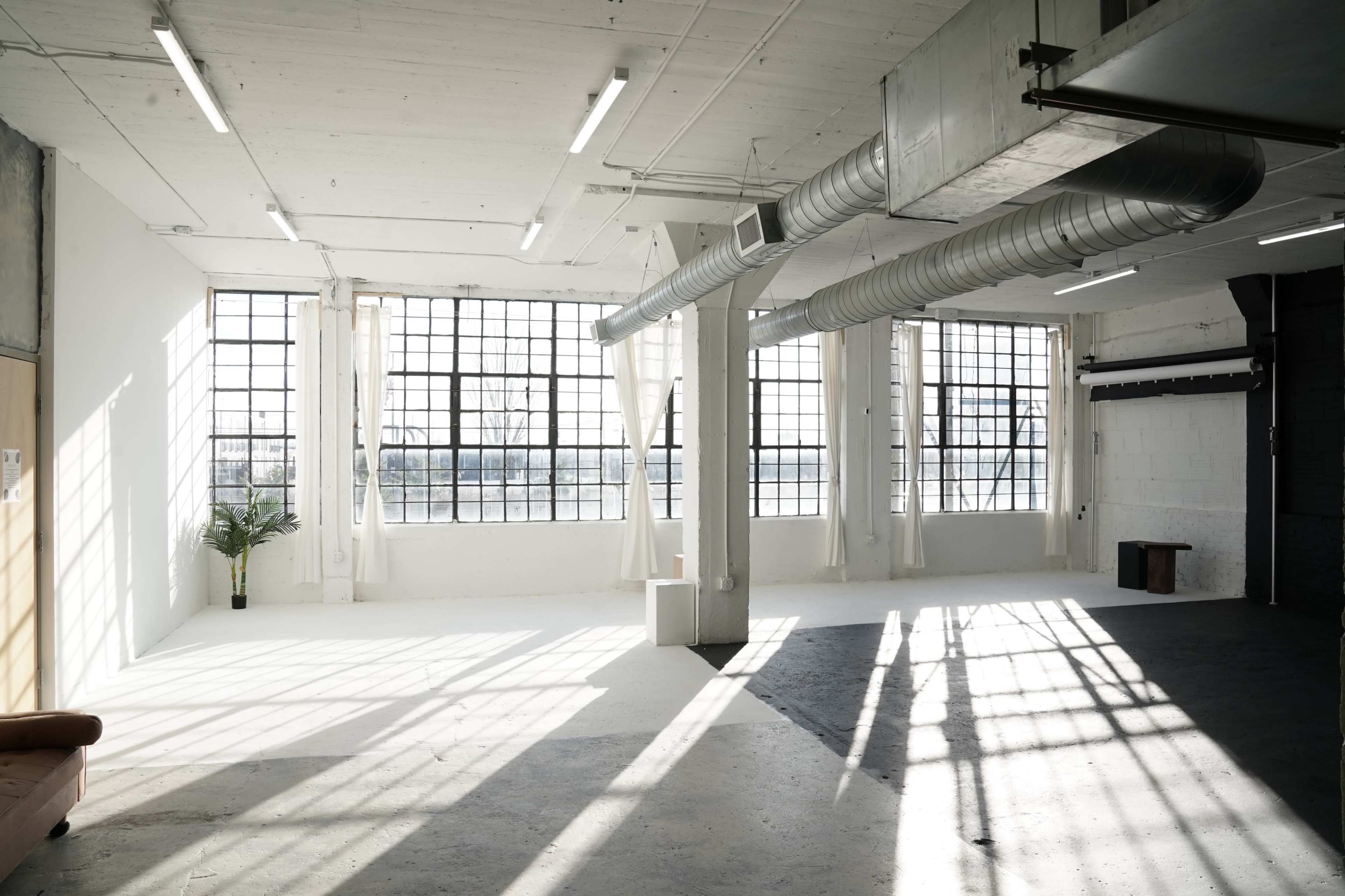 The image shows a spacious, modern interior with large windows allowing natural light to illuminate the white walls and concrete floor.
