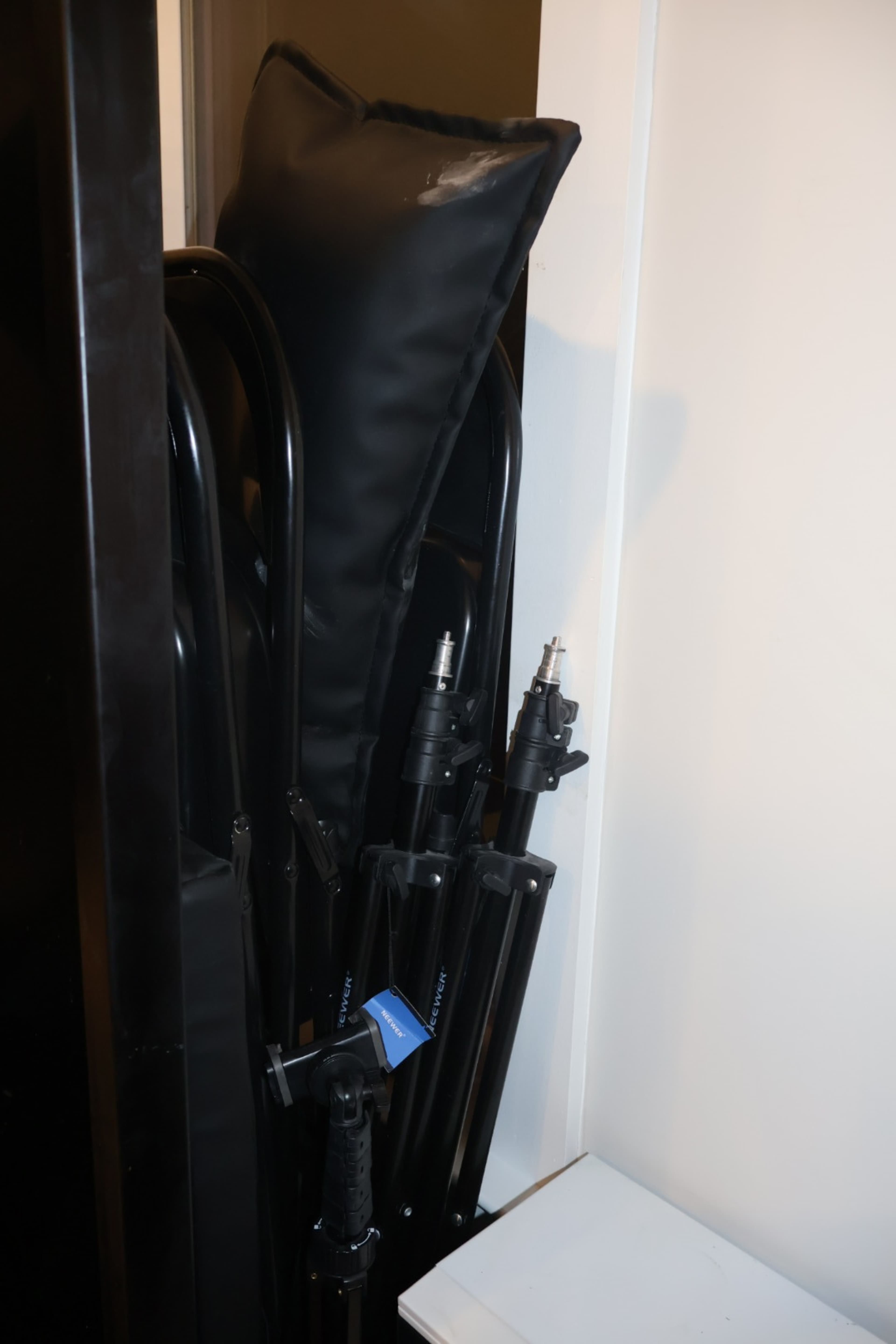 A black folding table and several black tripod stands are stored in a corner, partially obscured by a large, padded cushion.
