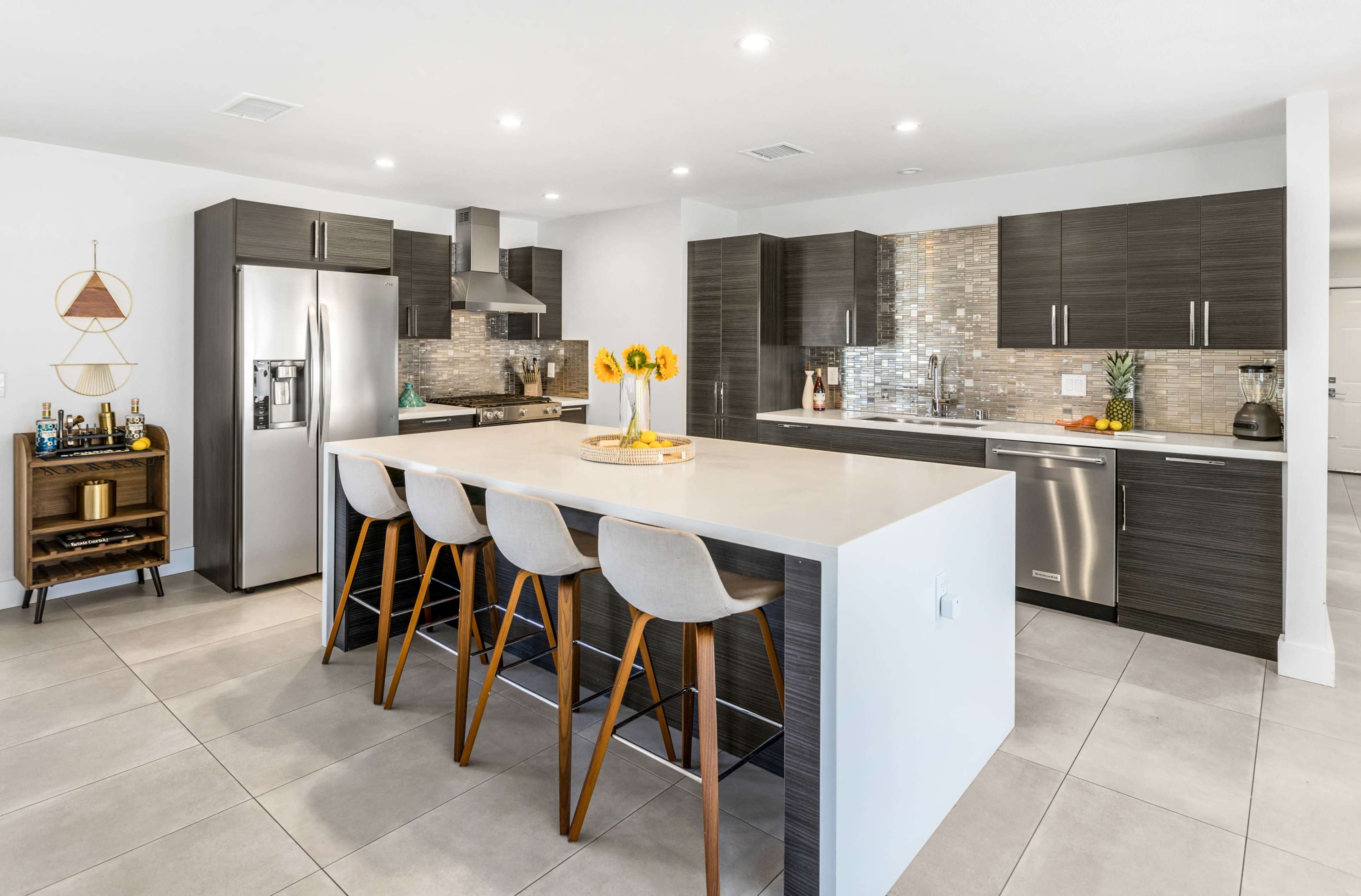 The image shows a modern kitchen with dark cabinetry, stainless steel appliances, and a large island with bar stools.