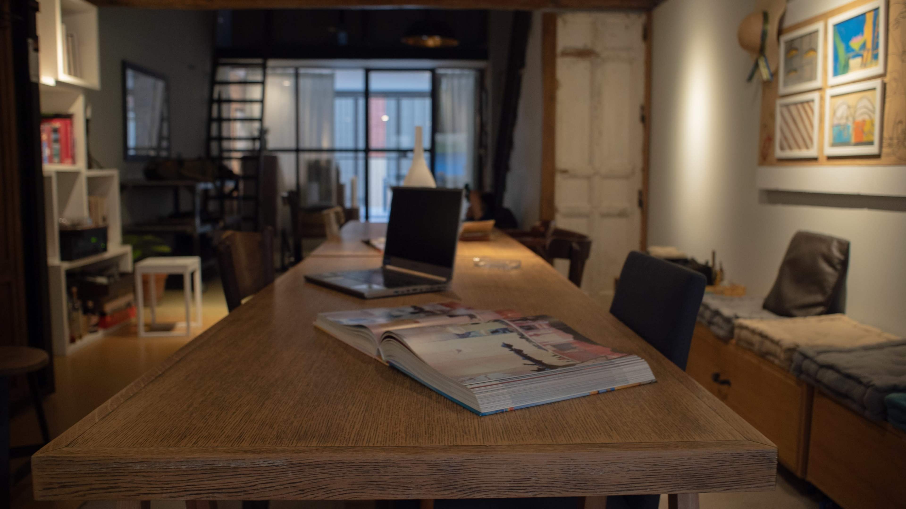 A long wooden table sits in a dimly lit room, with a laptop and an open magazine on its surface.