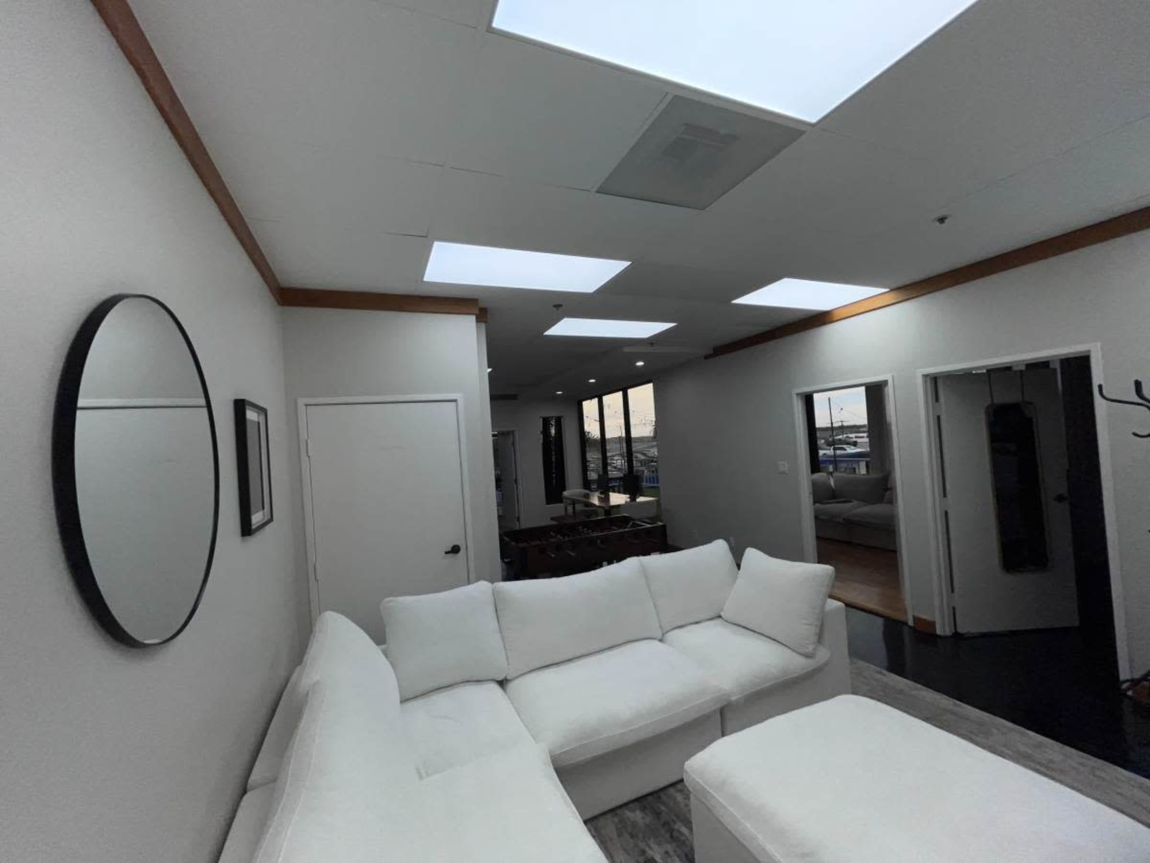 The image shows a bright interior space featuring a white sectional sofa, a round mirror on the wall, and multiple skylights illuminating the room.