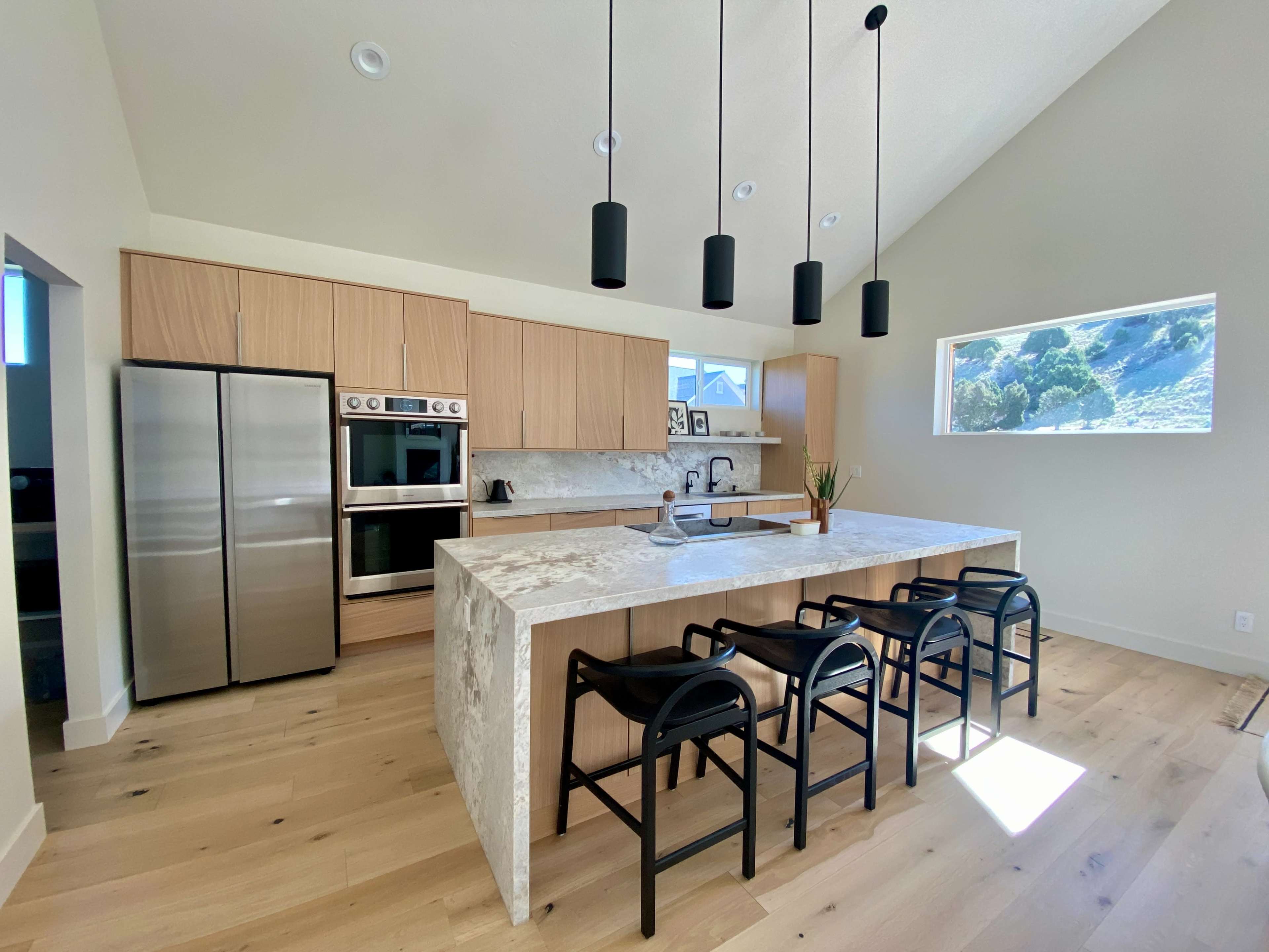 The image shows a modern kitchen with light wood cabinetry, stainless steel appliances, and a large island featuring bar stools.