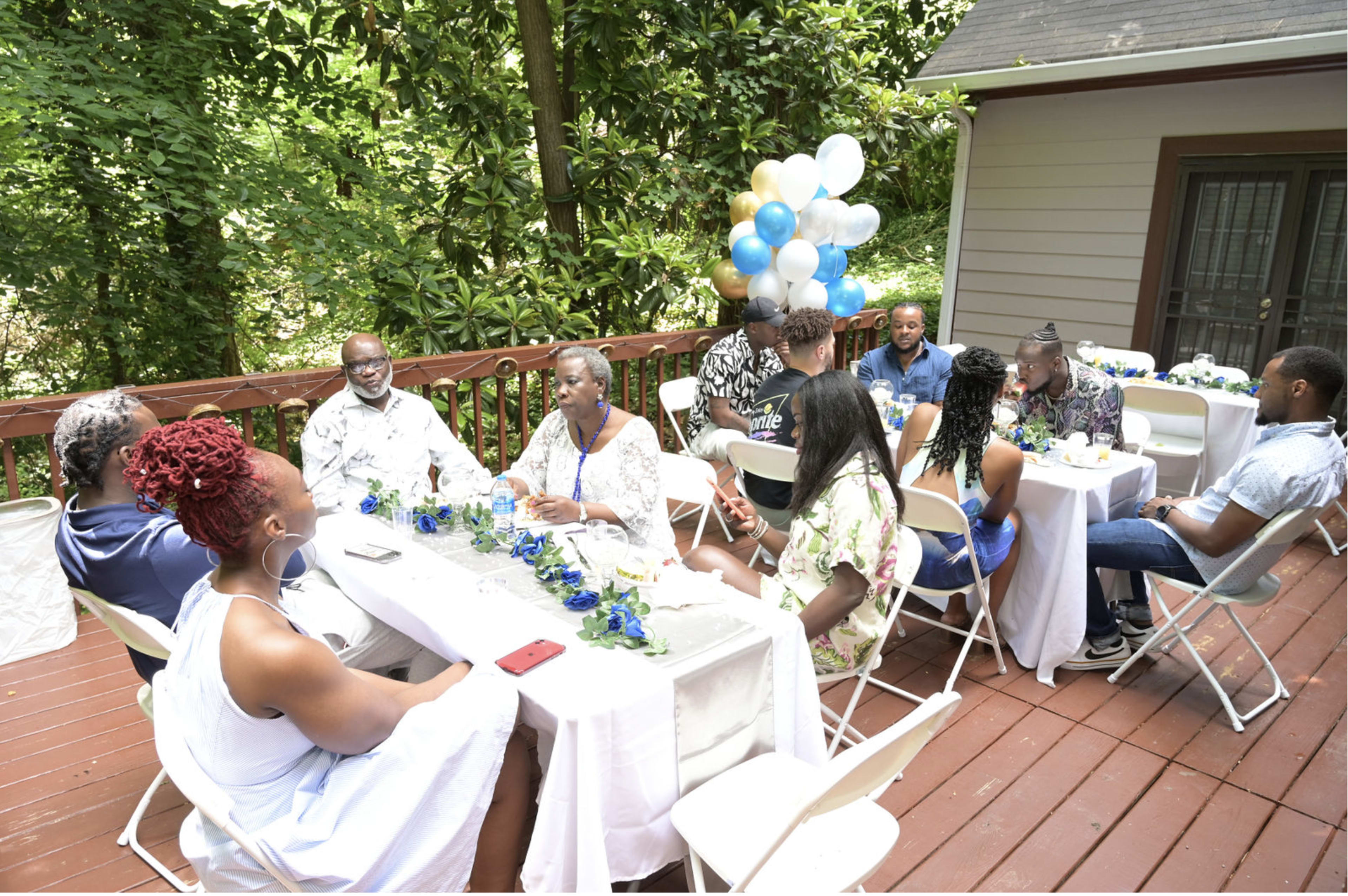 A group of people is gathered around a long table on a deck, enjoying a celebration amidst greenery and balloons.