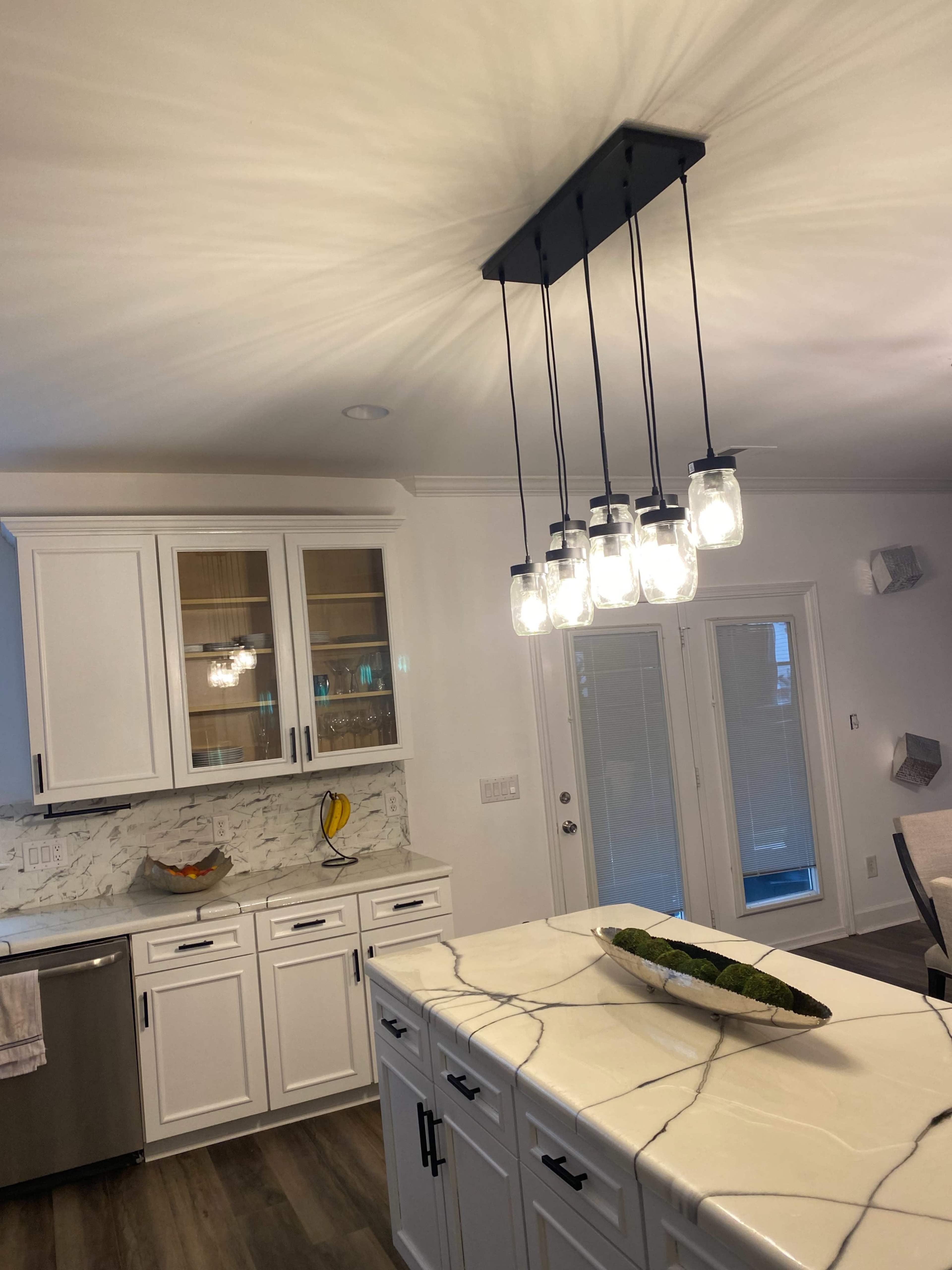 A modern kitchen features white cabinetry, a marble countertop with a centerpiece, and a pendant light with multiple glass bulbs hanging from the ceiling.