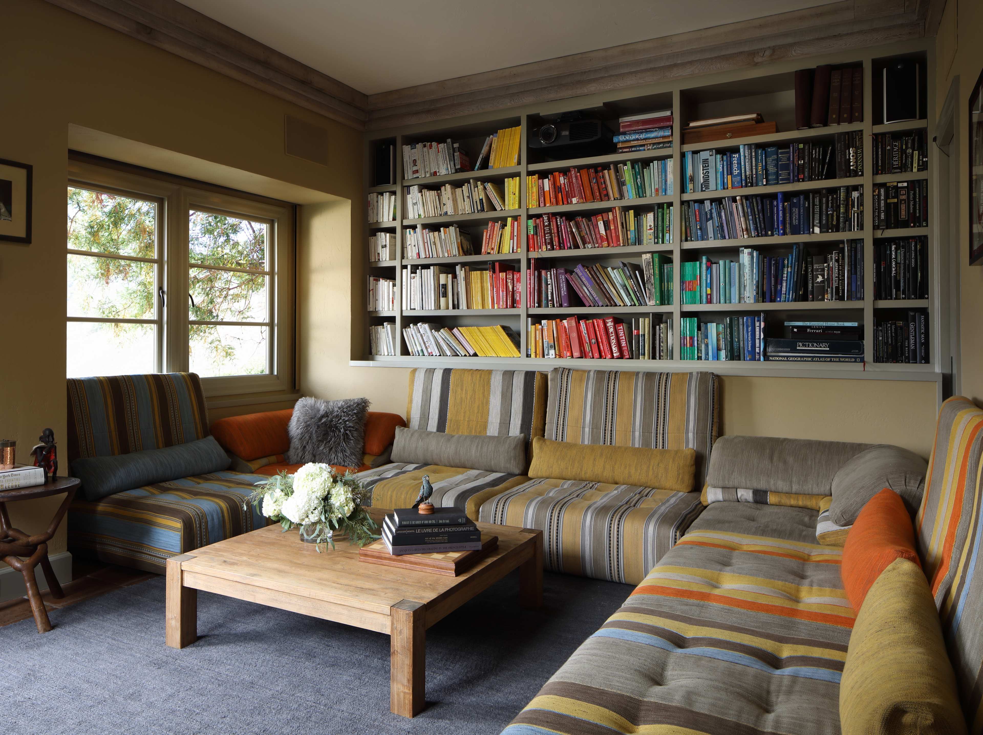 A cozy living room with a striped sectional sofa, a wooden coffee table, and a bookshelf filled with colorful books.