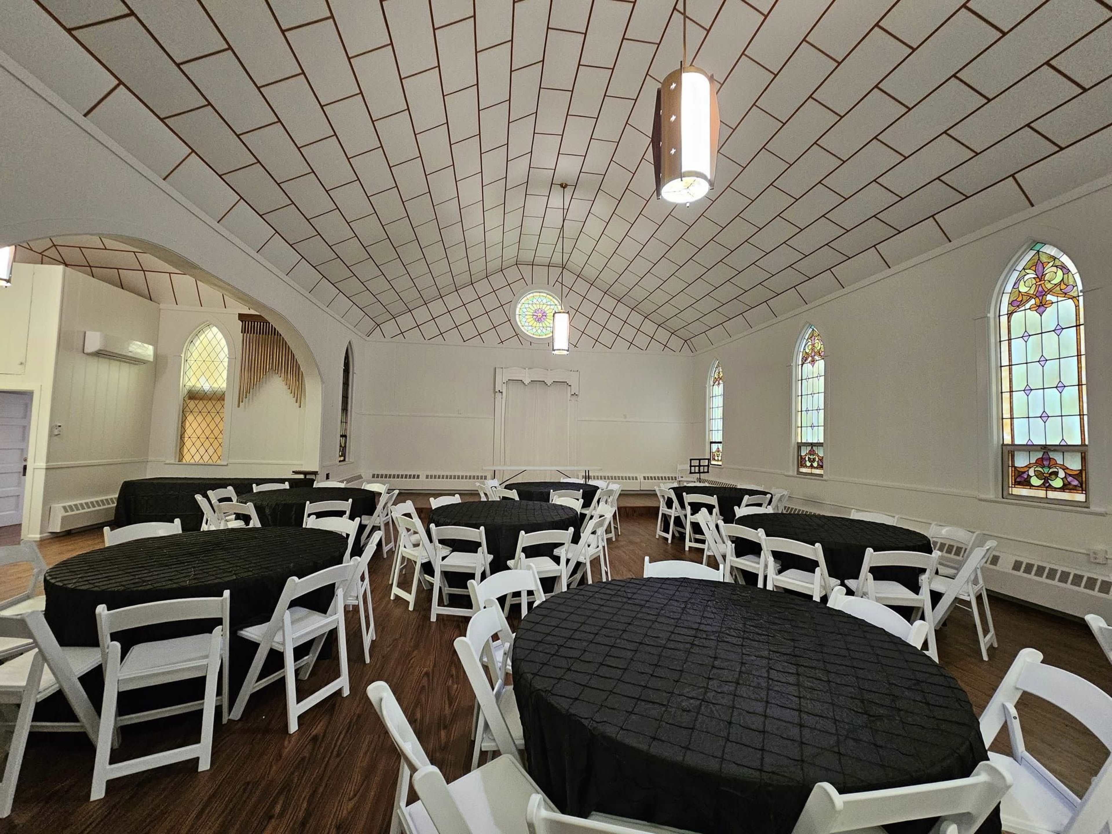 A spacious event hall with round tables covered in black cloth and stained glass windows lining the walls.